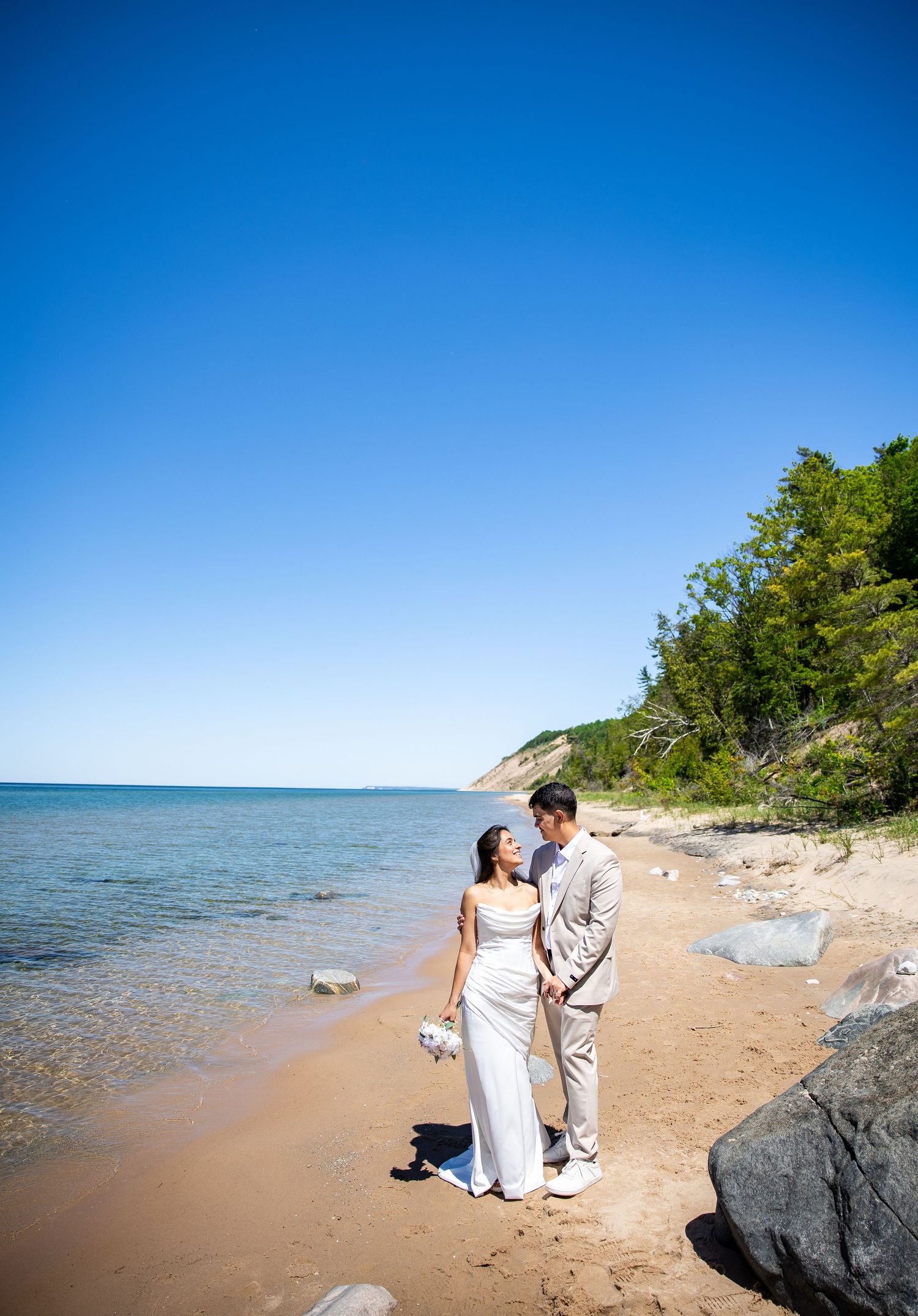 Bride in a white gown and groom in a tan suit embrace on a sandy lakeshore beach beneath a clear blue sky with wooded dunes nearby.