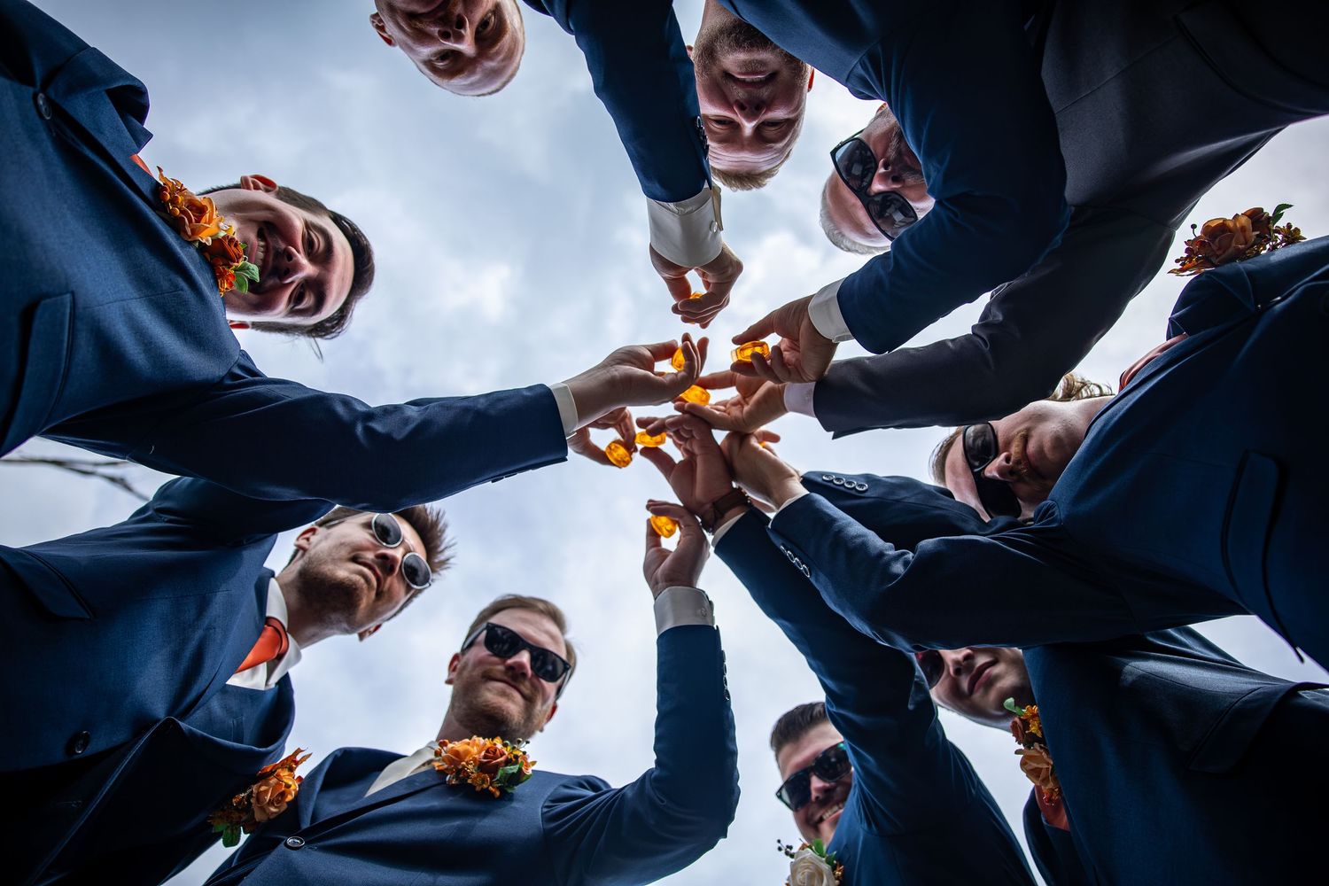 Low-angle view of groomsmen in navy suits and orange floral boutonnieres clinking drinks in a circle under a bright sky.