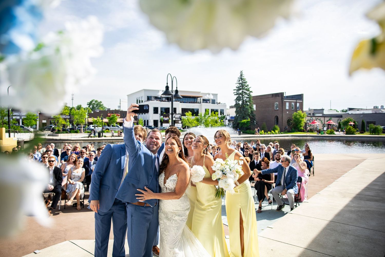 Bride, bridesmaids and groomsmen pose for a selfie at an outdoor waterfront wedding ceremony while seated guests look on.