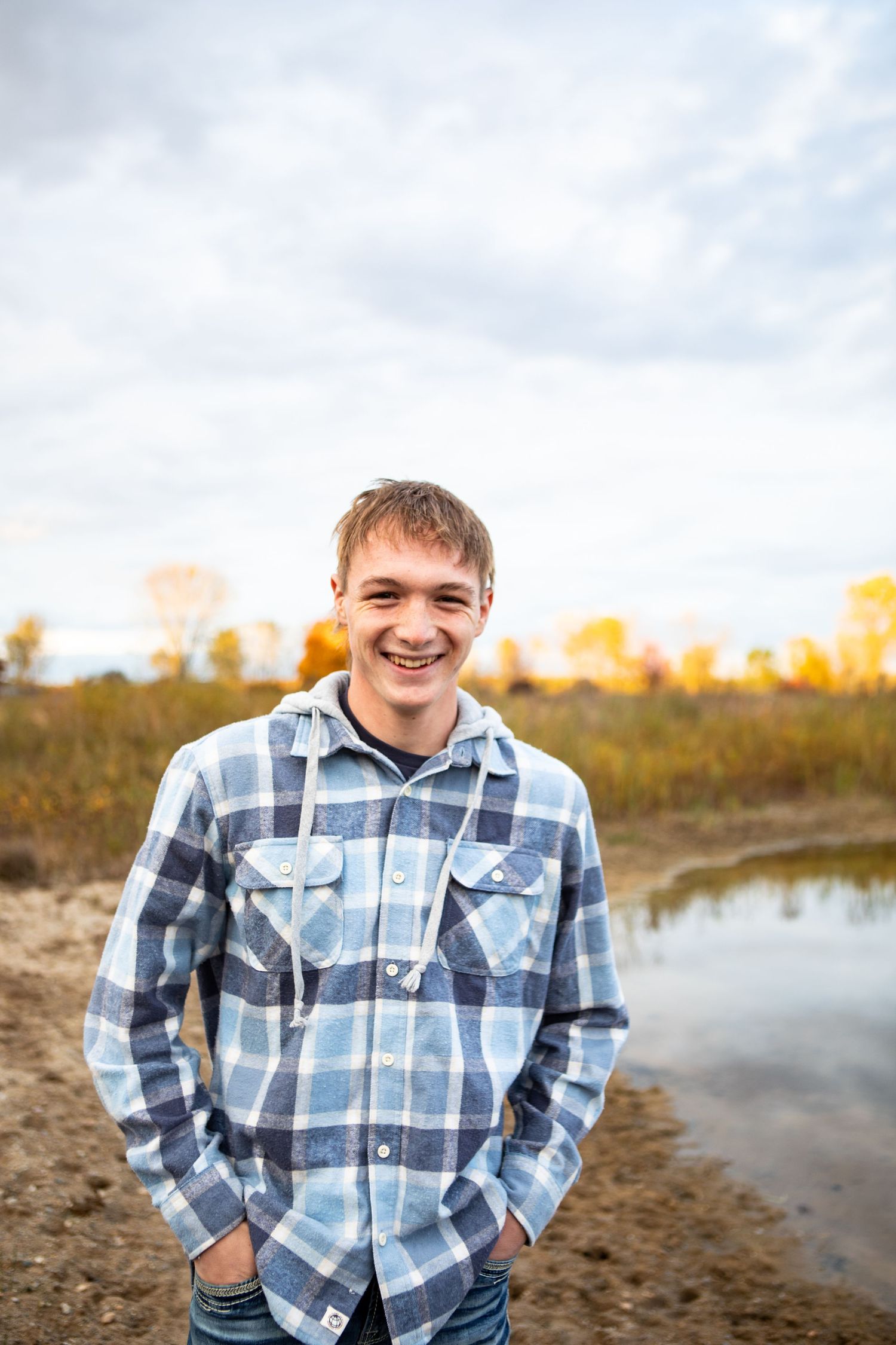 A person wearing a plaid button-up shirt smiles while standing near a lake during golden hour.