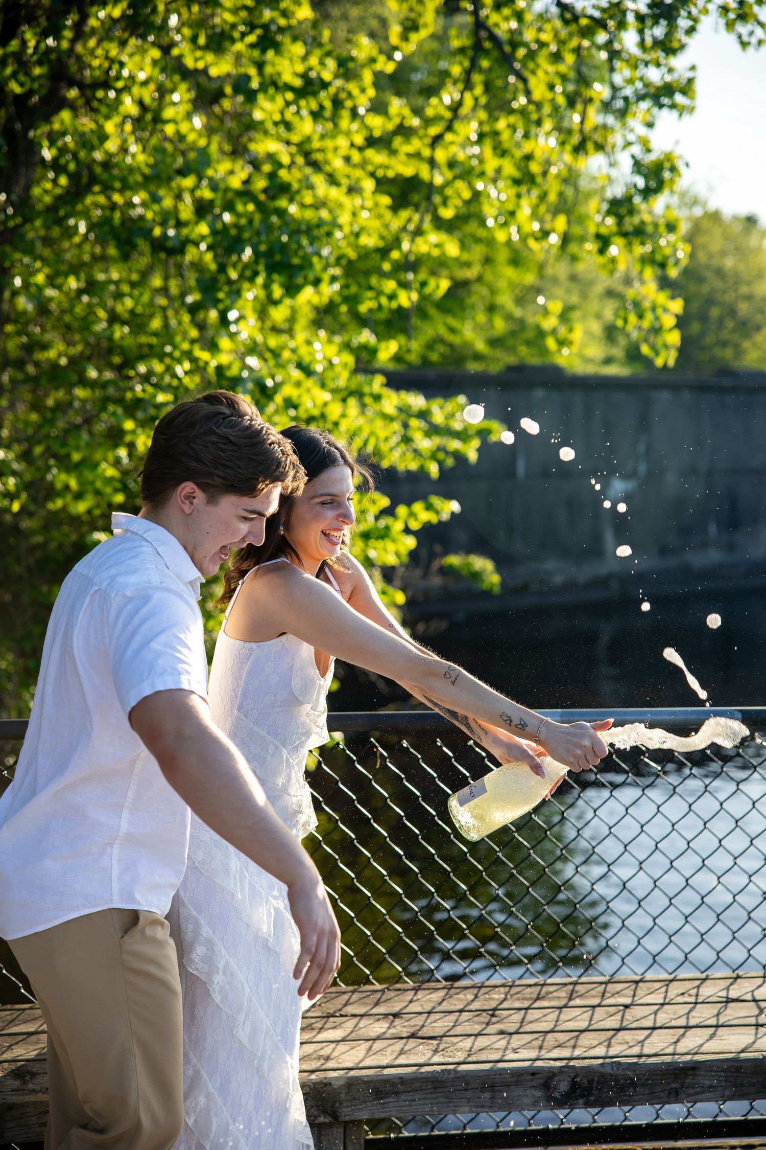 Happy couple pouring champagne over a bridge railing surrounded by lush green trees.