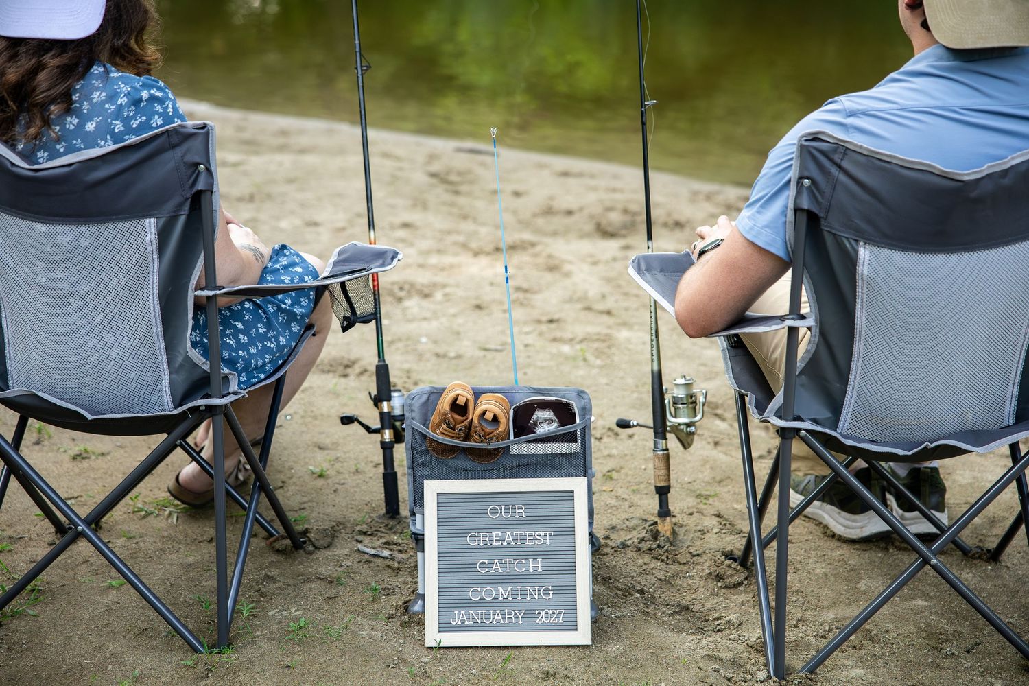 Couple fishing by a lake with a letter board sign reading Our Greatest Catch Coming Soon for a pregnancy announcement.