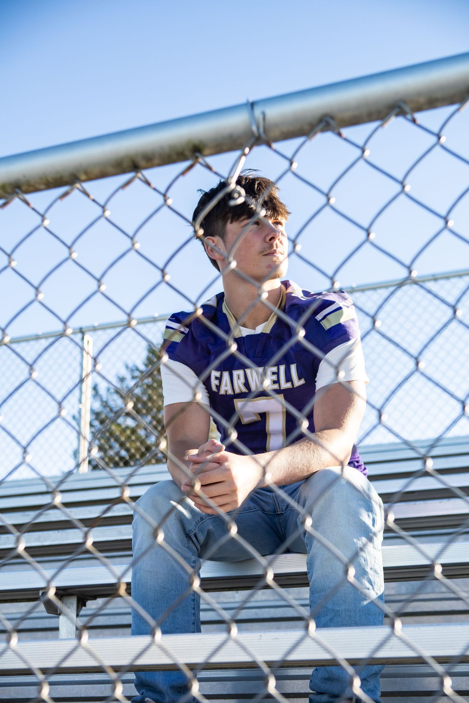 Young man in Farwell football jersey and helmet sits on bleachers behind chain-link fence on sunny day.