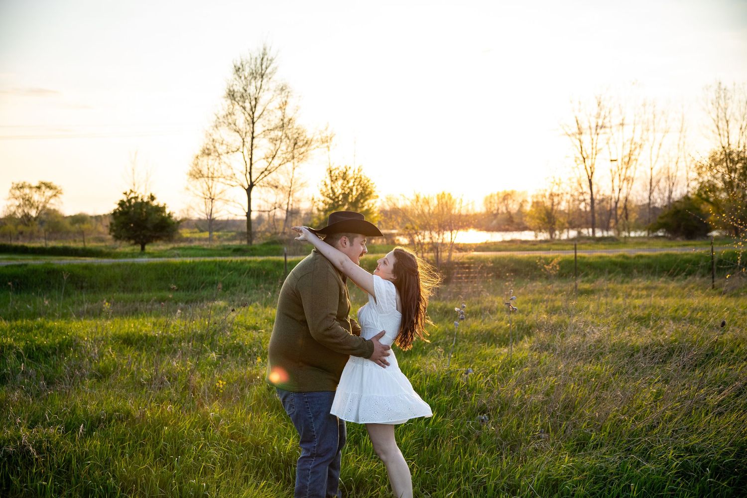 Couple sharing a romantic kiss at golden hour in a lush green field with trees and glowing sunset backdrop.