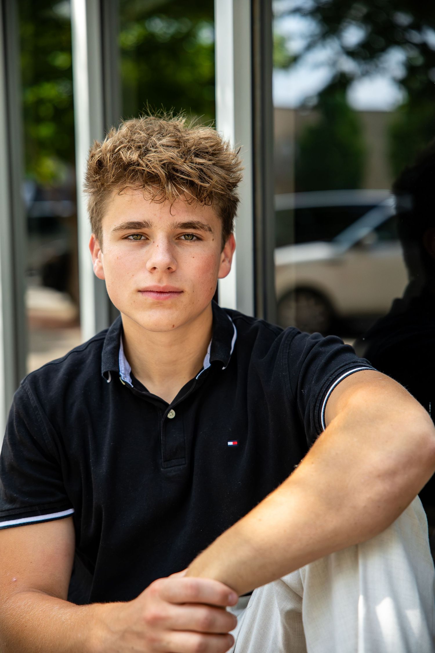 Young man in navy Tommy Hilfiger polo sitting outdoors, posing confidently with arms crossed.