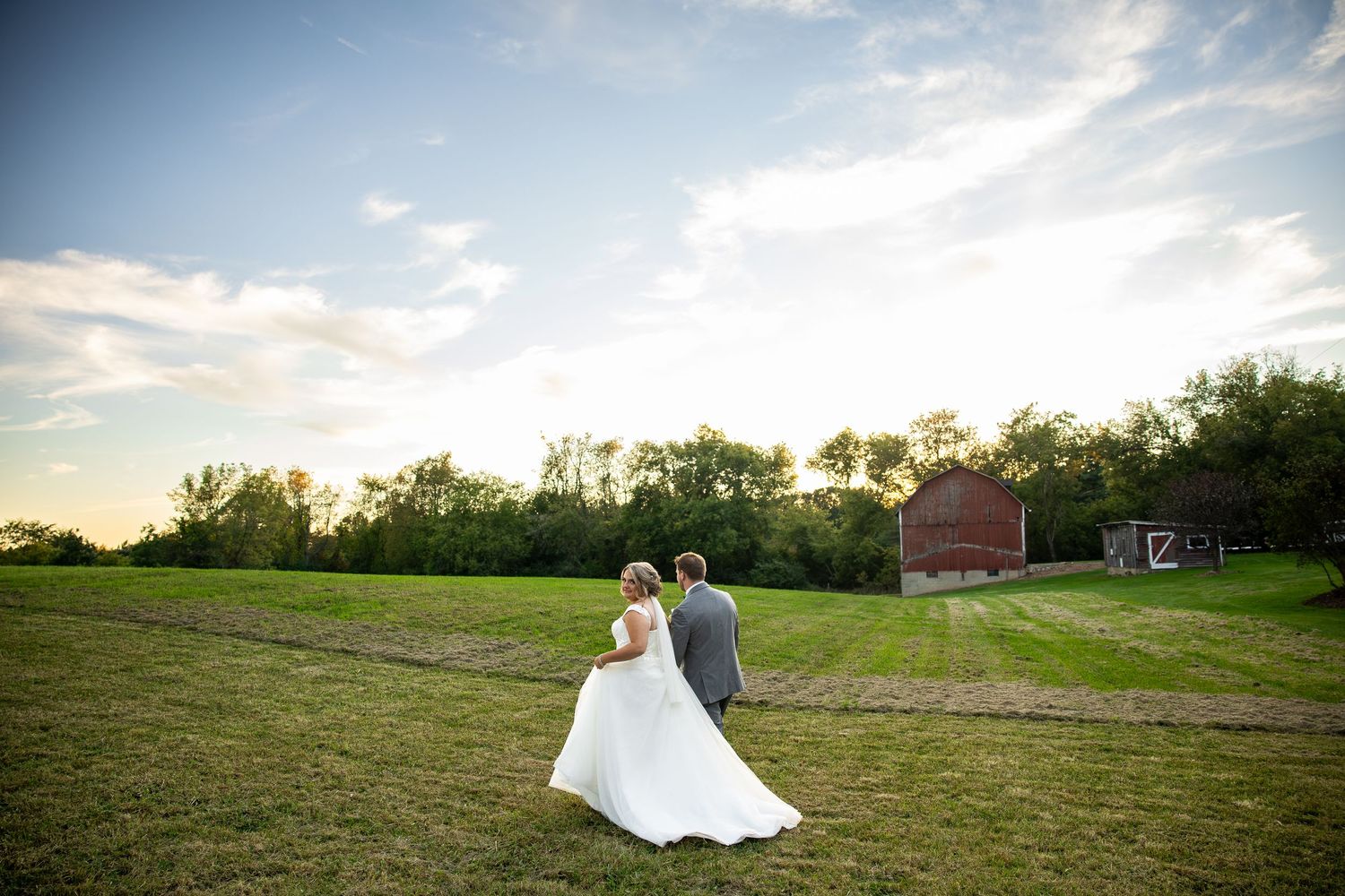 Bride in a white gown and groom in a gray suit walk across a grassy field toward a red barn at sunset during a rustic wedding.