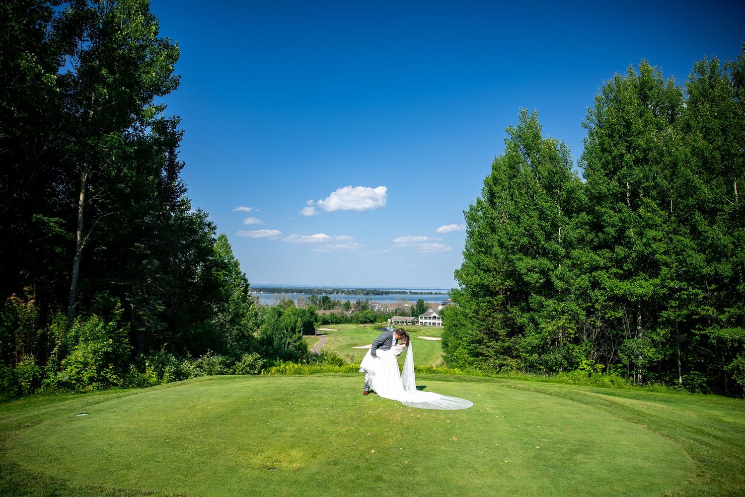 Bride in a long white wedding gown kneels on a manicured golf course green framed by tall evergreen trees with a lake and blue sky behind.