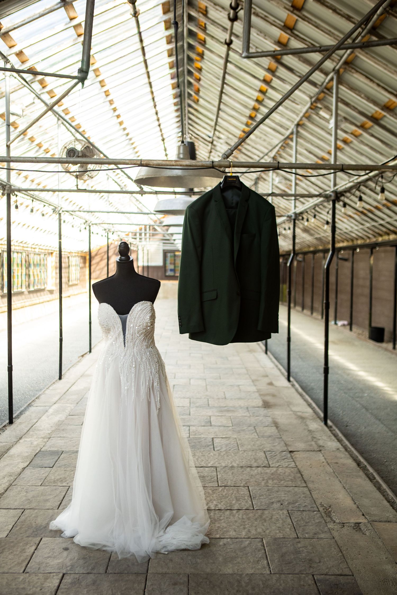 Wedding dress on mannequin and dark green groom suit jacket hanging in elegant greenhouse venue.