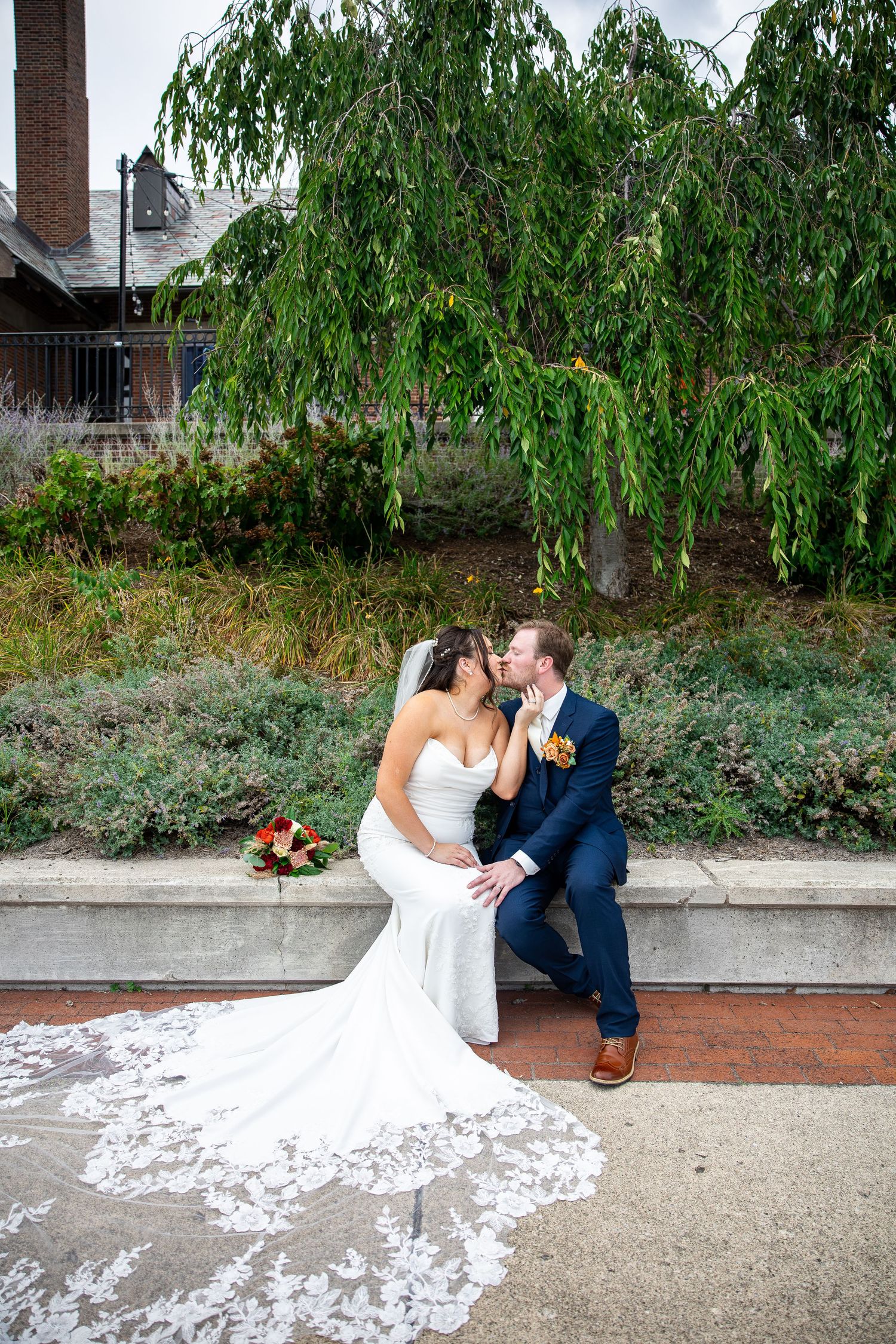 Bride in a white lace wedding gown and groom in a navy suit share a kiss on a stone ledge beneath a weeping tree.