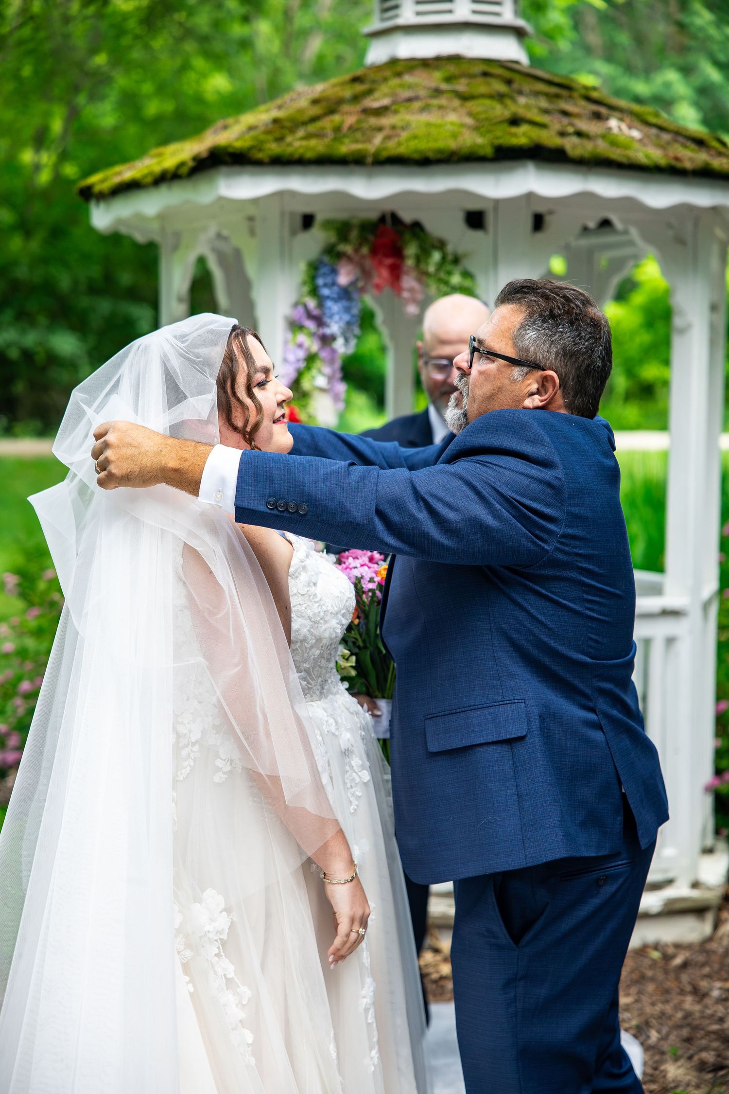 Groom in blue suit lifting bride's veil during outdoor wedding ceremony at white gazebo.