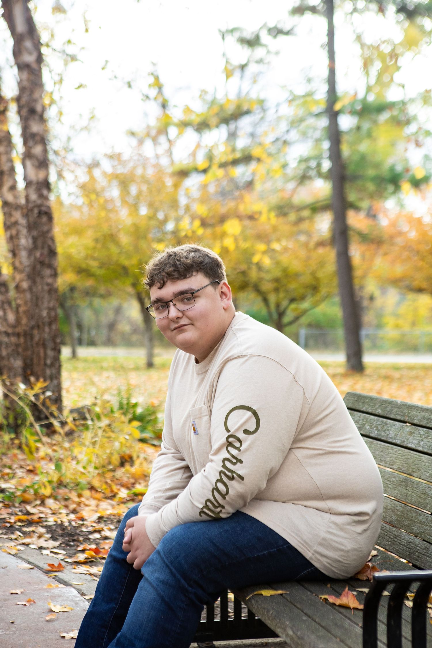 A person in a cream-colored sweatshirt sits thoughtfully on a wooden bench surrounded by autumn leaves and trees.