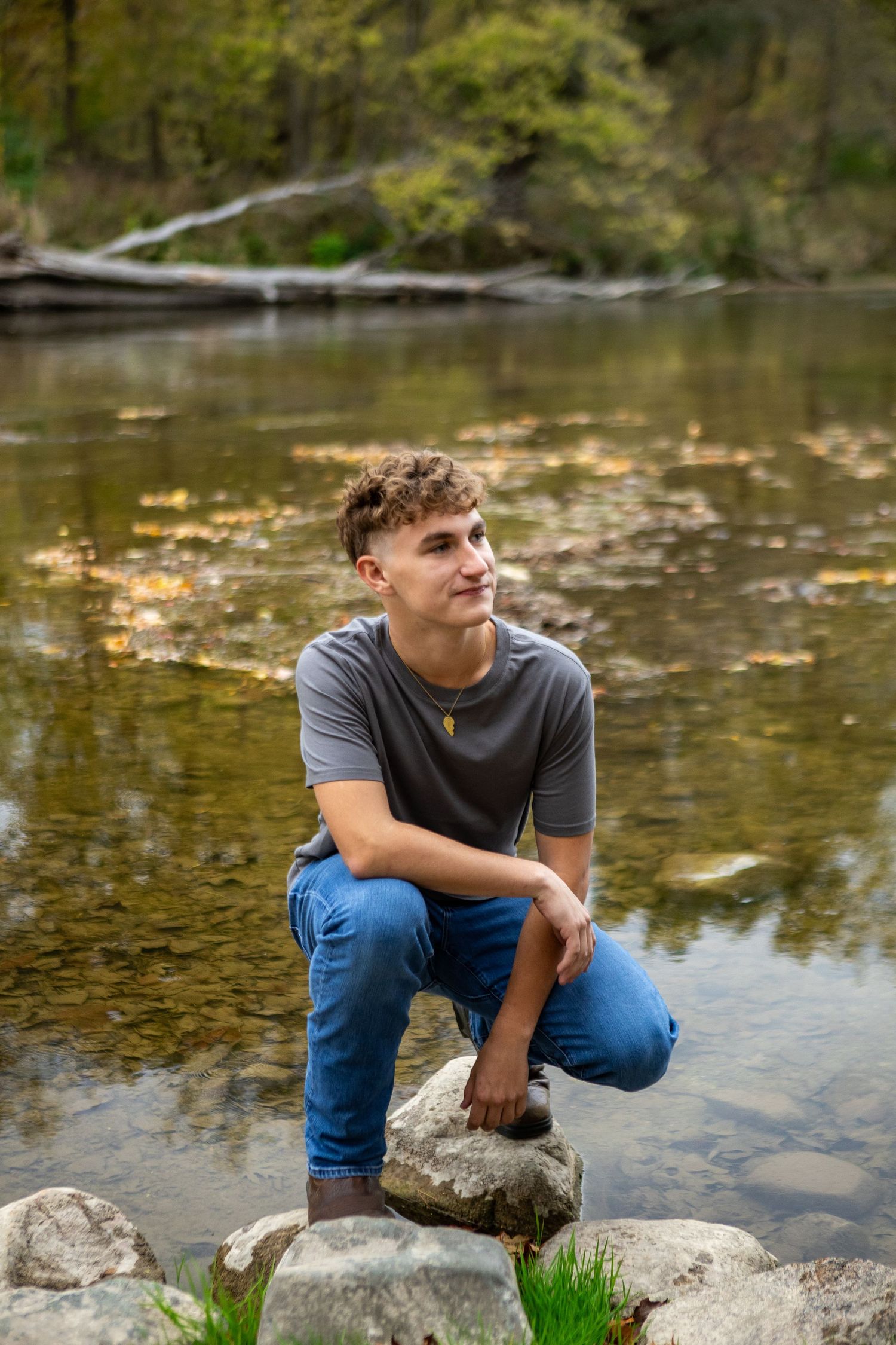 A person in a gray t-shirt and blue jeans sits on rocks beside a tranquil stream in nature.