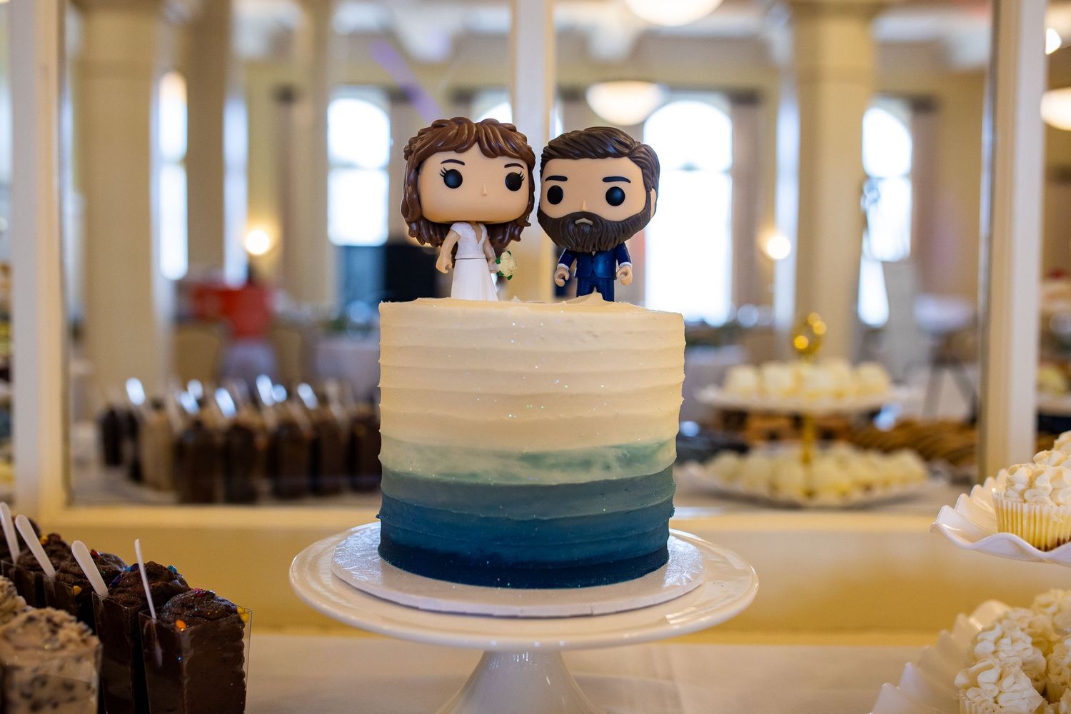 Wedding cake with ombre blue design topped with Funko Pop bride and groom figurines on a white cake stand.