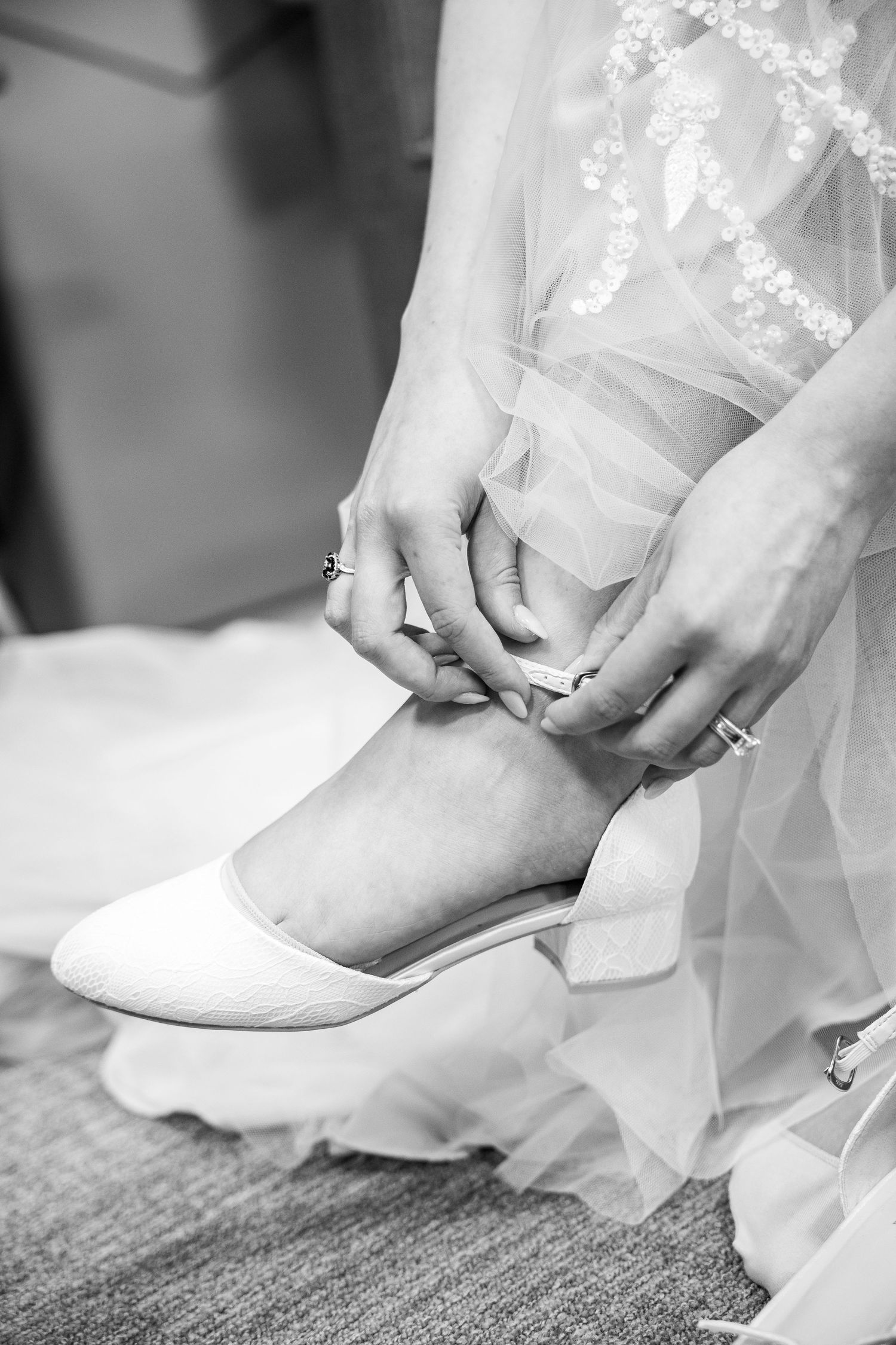 Bride putting on white wedding shoes, adjusting ankle strap, wearing lace gown, black and white photo.
