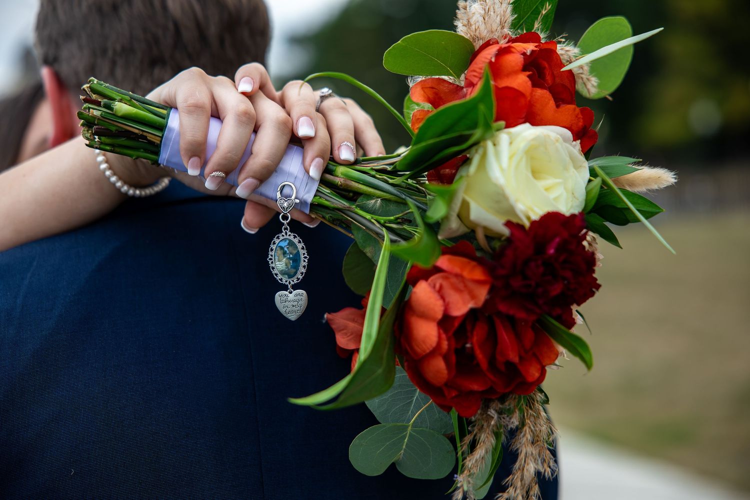 Bride's hands hold a bouquet of orange and cream roses with green leaves, wrapped in ribbon with a heart and photo charm, over a dark suit.