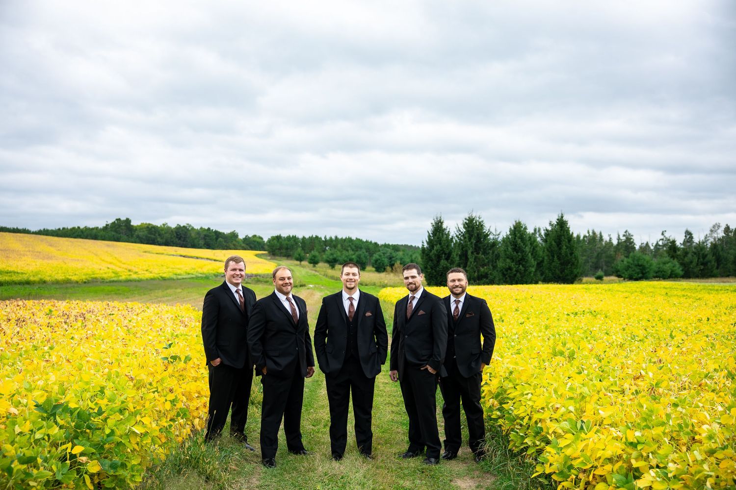 Five groomsmen in black suits stand together on a grass path between blooming yellow fields under a cloudy sky with evergreen trees behind.