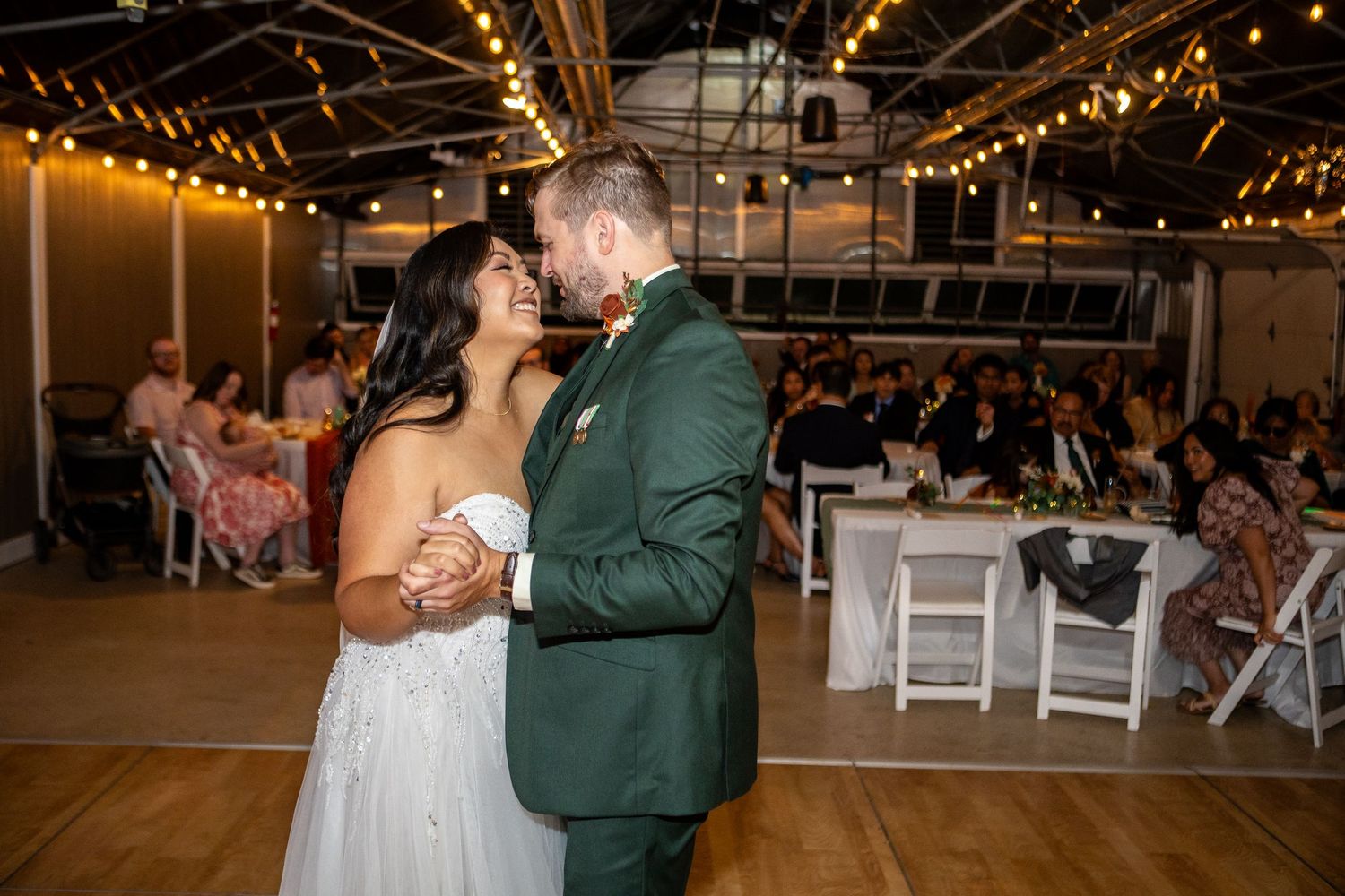 Bride and groom share their first dance at a wedding reception with string lights and guests seated in the background.