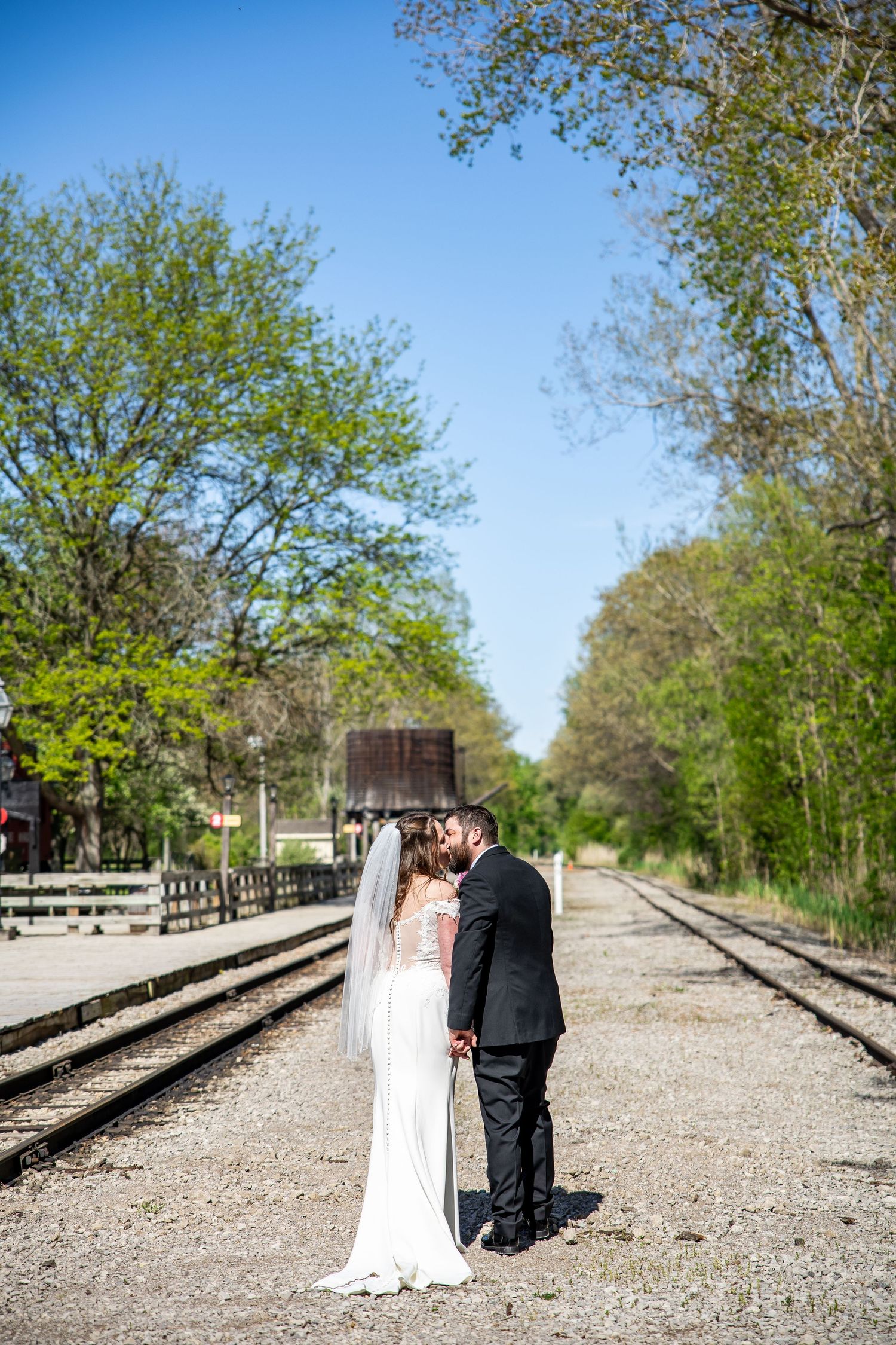 Bride and groom kissing on railroad tracks surrounded by lush green trees under a clear blue sky.