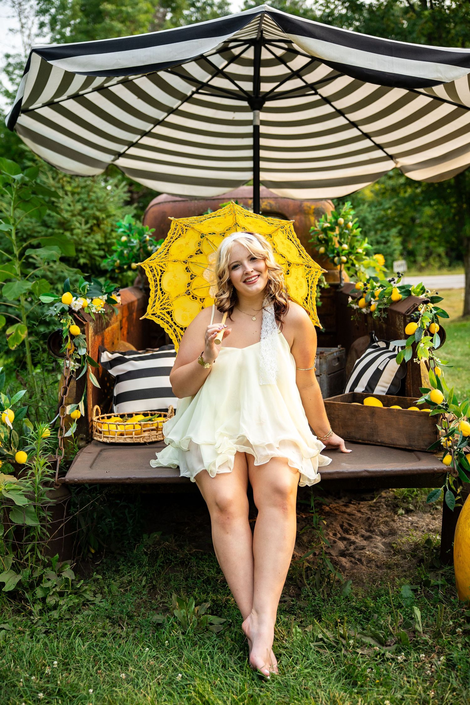 Smiling blonde woman in white dress holding yellow lace umbrella, sitting on rustic bench with lemons and striped parasol outdoors.