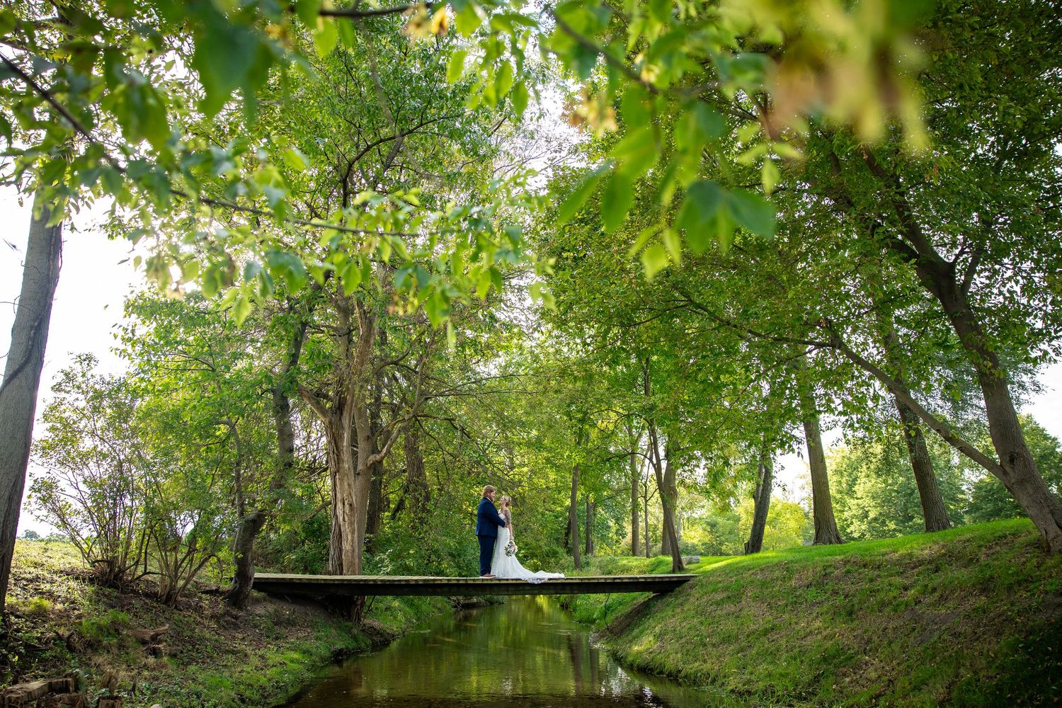 Bride and groom embrace on a wooden footbridge over a calm stream, surrounded by lush green trees in a sunlit park.