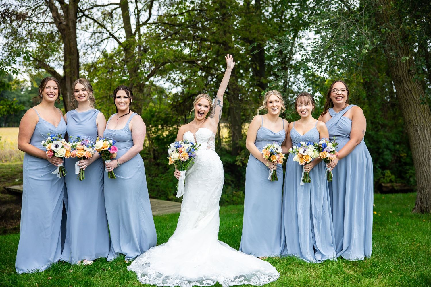 Bride in a white lace gown raises her arm in joy, surrounded by six bridesmaids in dusty blue dresses holding bouquets outdoors.