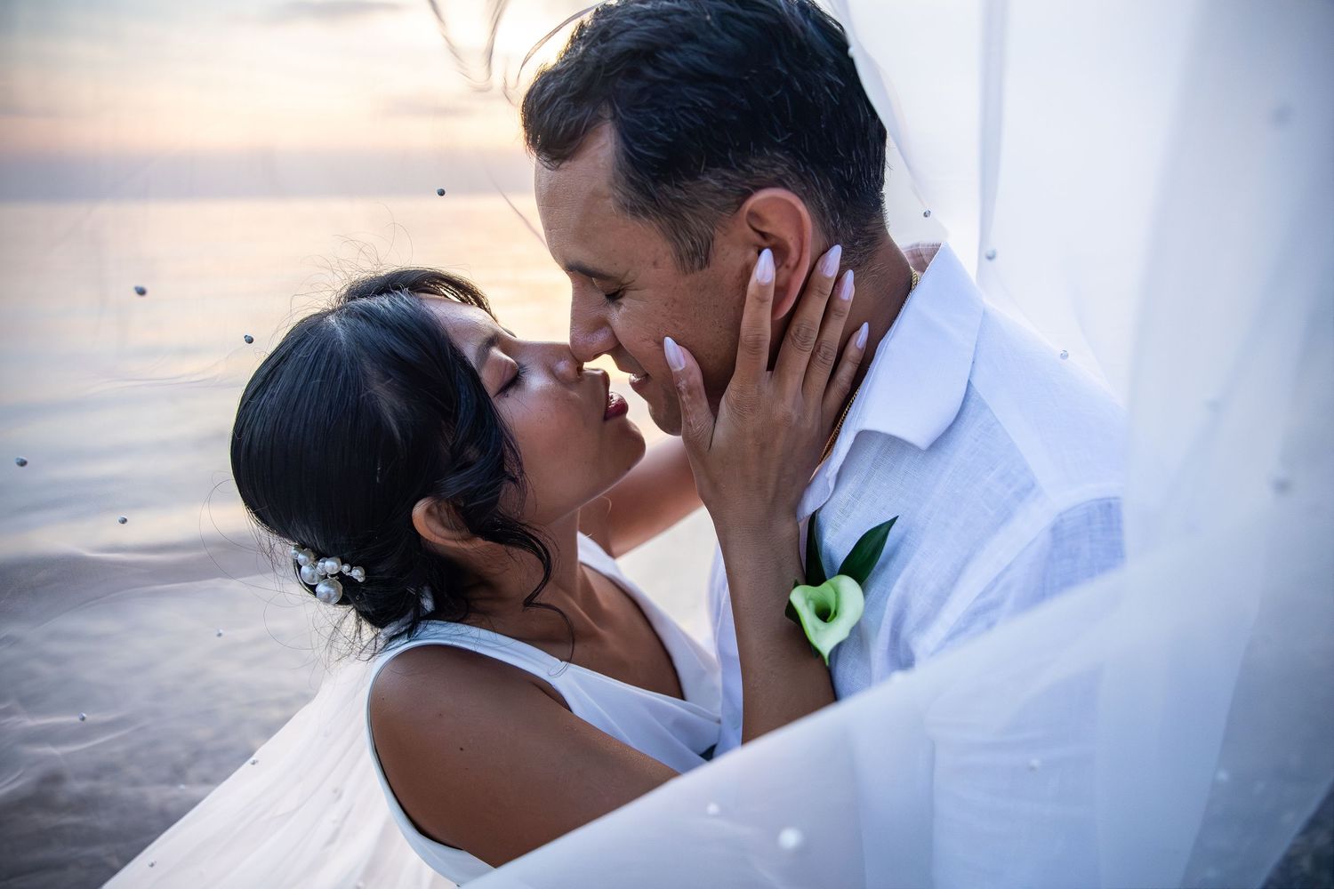Couple sharing a romantic kiss at sunset on a beach during their wedding ceremony, dressed in white attire.