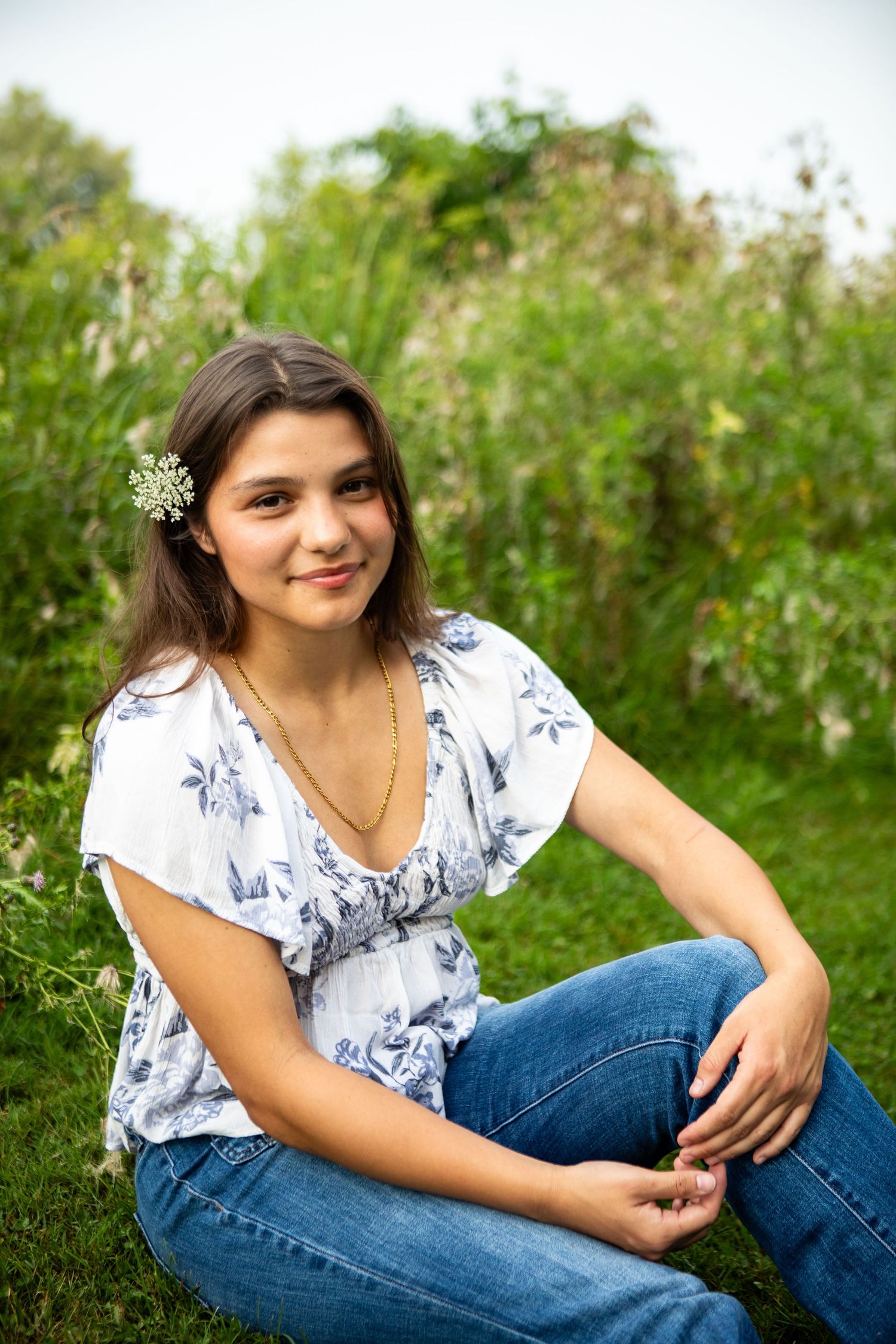 A person in a white floral top and blue jeans poses casually on grass with wildflowers in the background.