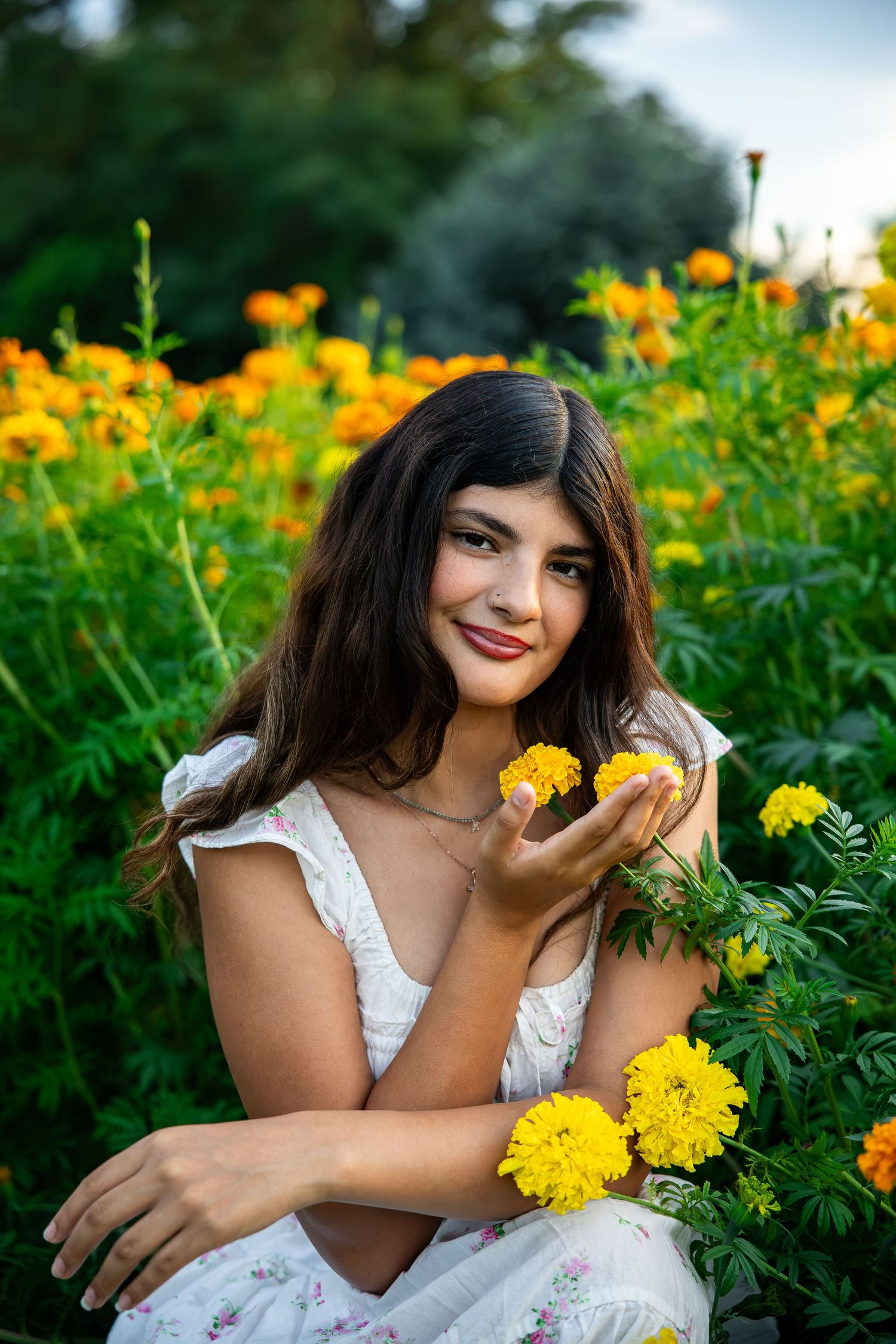 Young woman in white floral dress holding yellow marigolds while sitting in a vibrant flower garden.