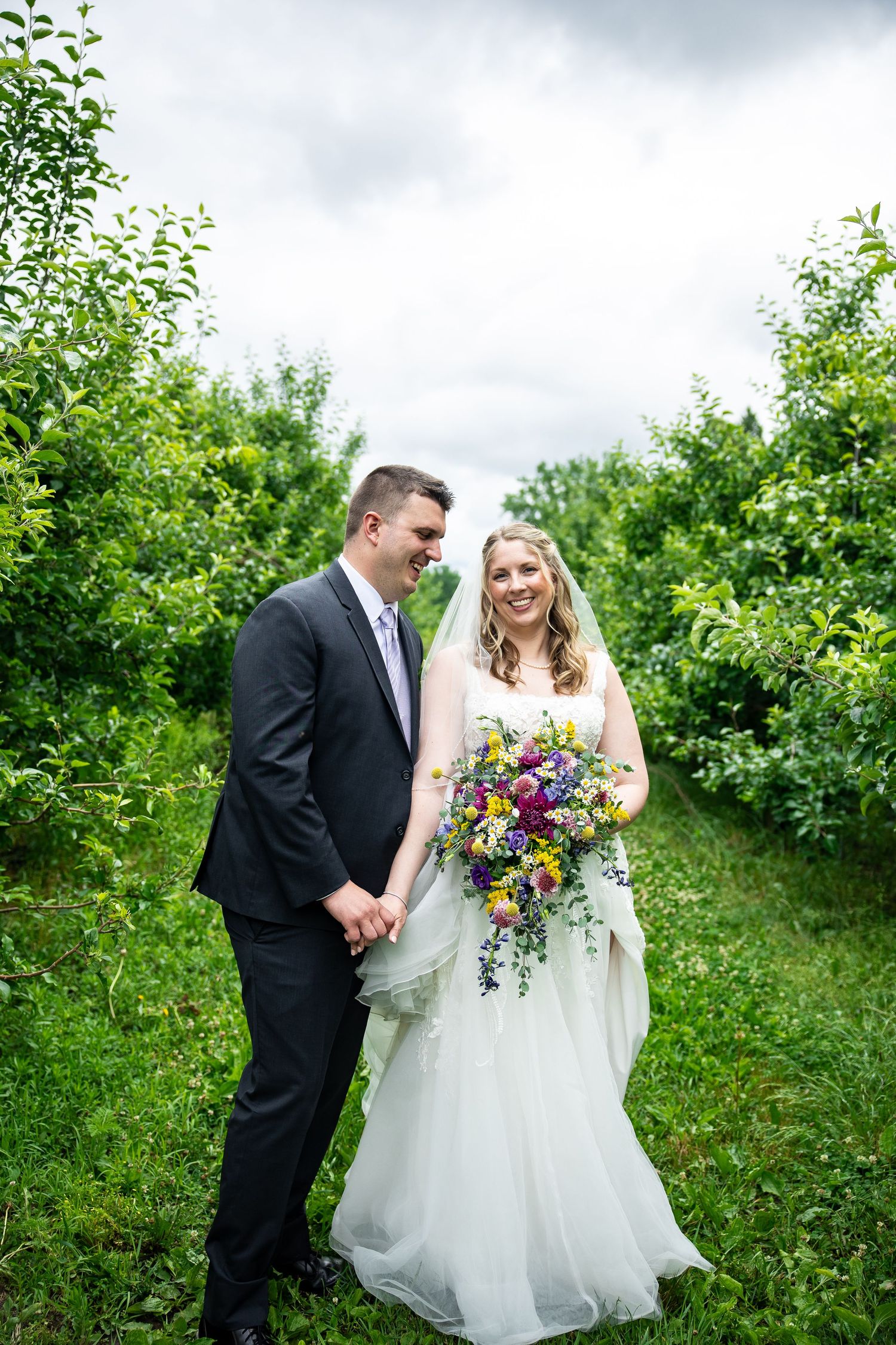Bride and groom hold hands smiling in a lush green orchard, bride holding colorful wildflower bouquet in white gown.