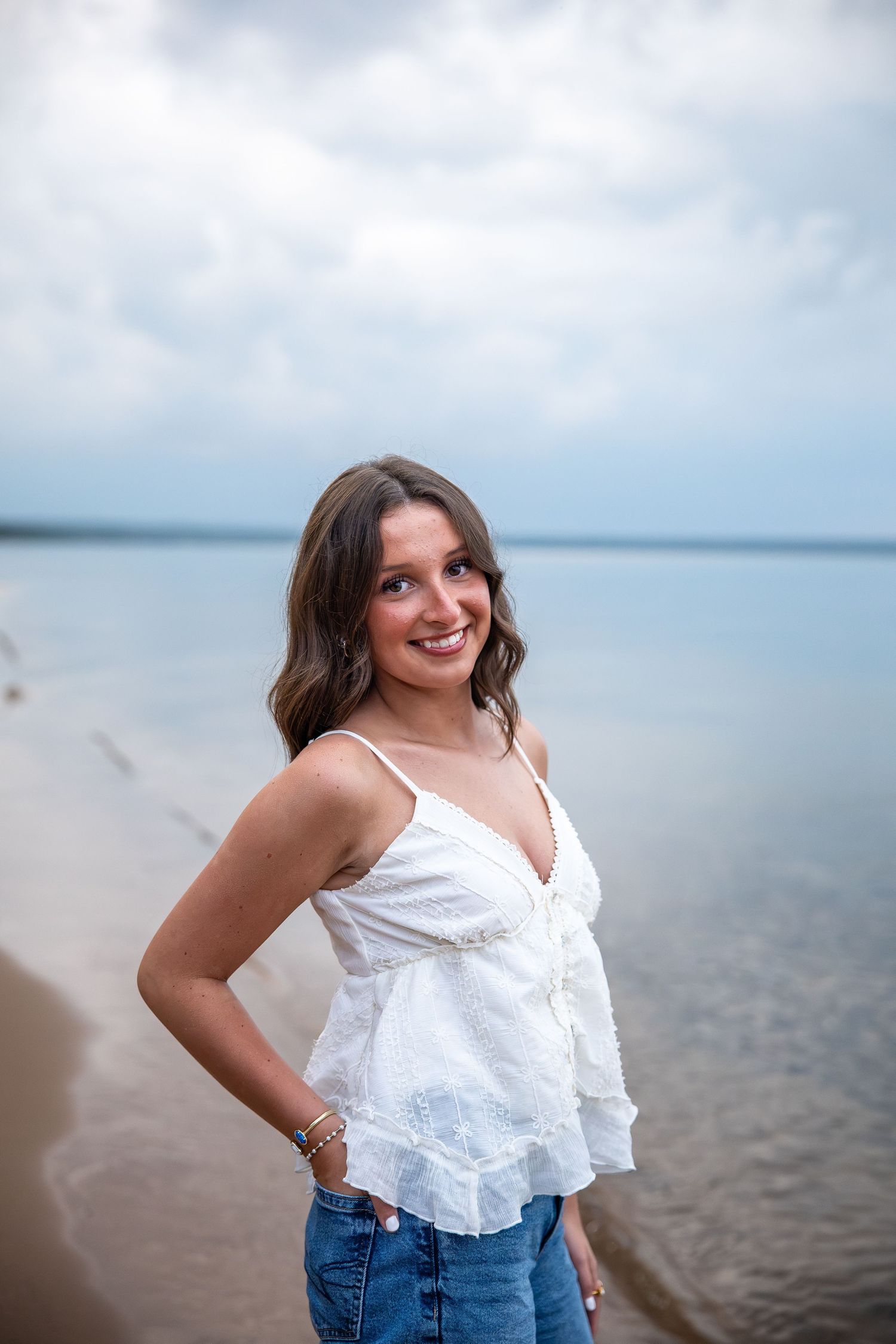 Young woman in white top and jeans smiling on a beach with calm water and cloudy sky behind her.