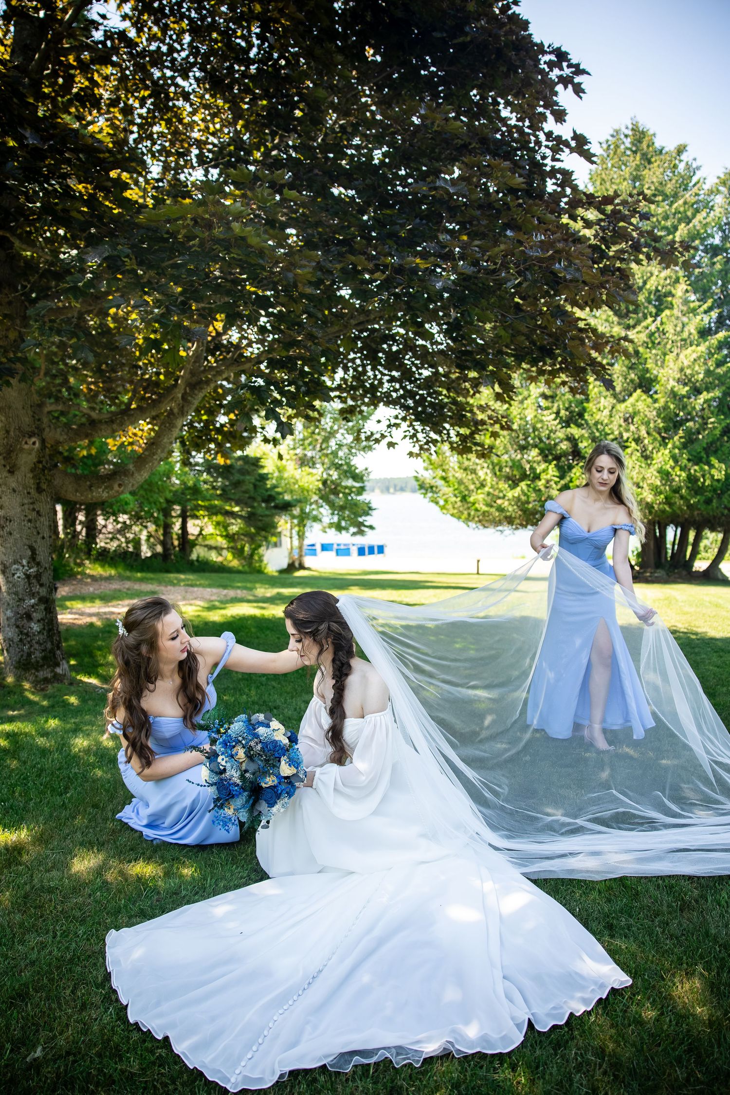 Bride in white gown sits on grass with two bridesmaids in blue dresses holding her veil outdoors.
