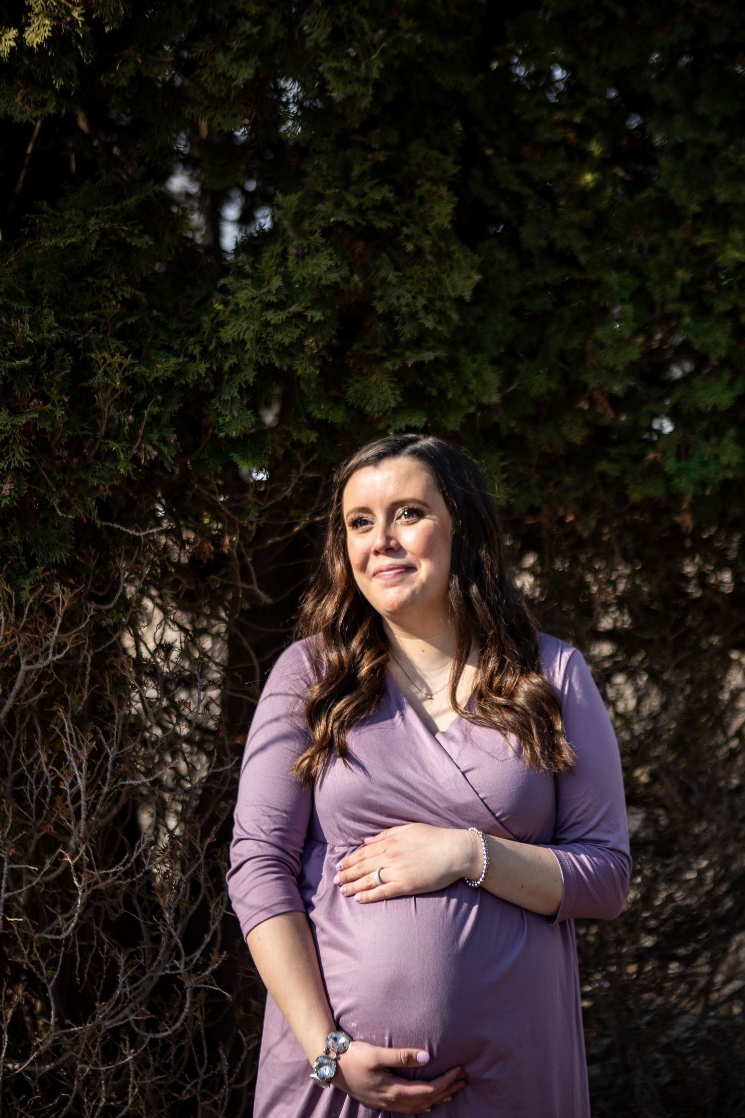 Pregnant woman in lavender wrap dress cradling baby bump, smiling outdoors with lush green trees behind her.