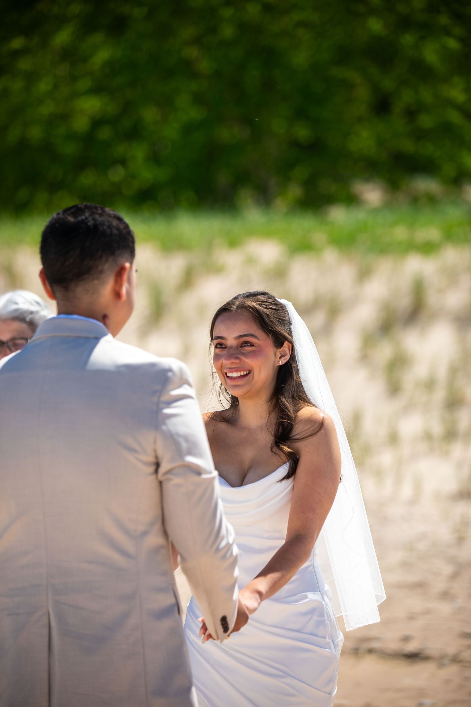Smiling bride in white gown and veil holds hands with groom in light suit during outdoor beach wedding ceremony.
