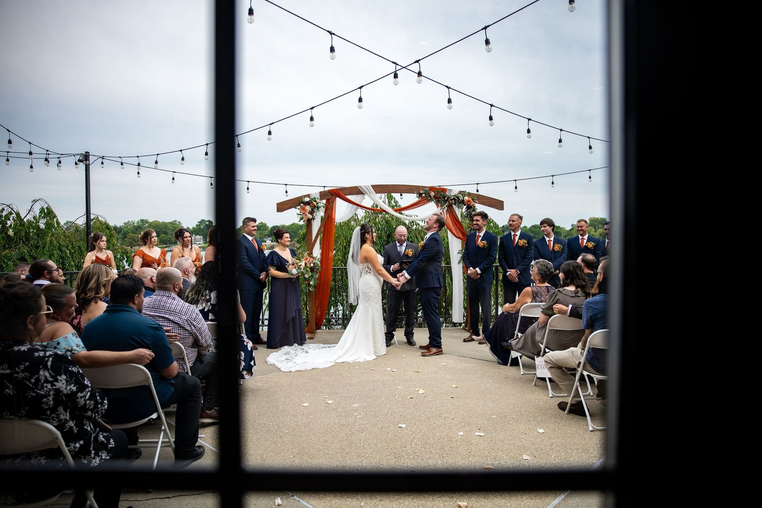 Bride and groom exchange vows under a rustic wooden arch at an outdoor wedding ceremony as seated guests look on beneath string lights.