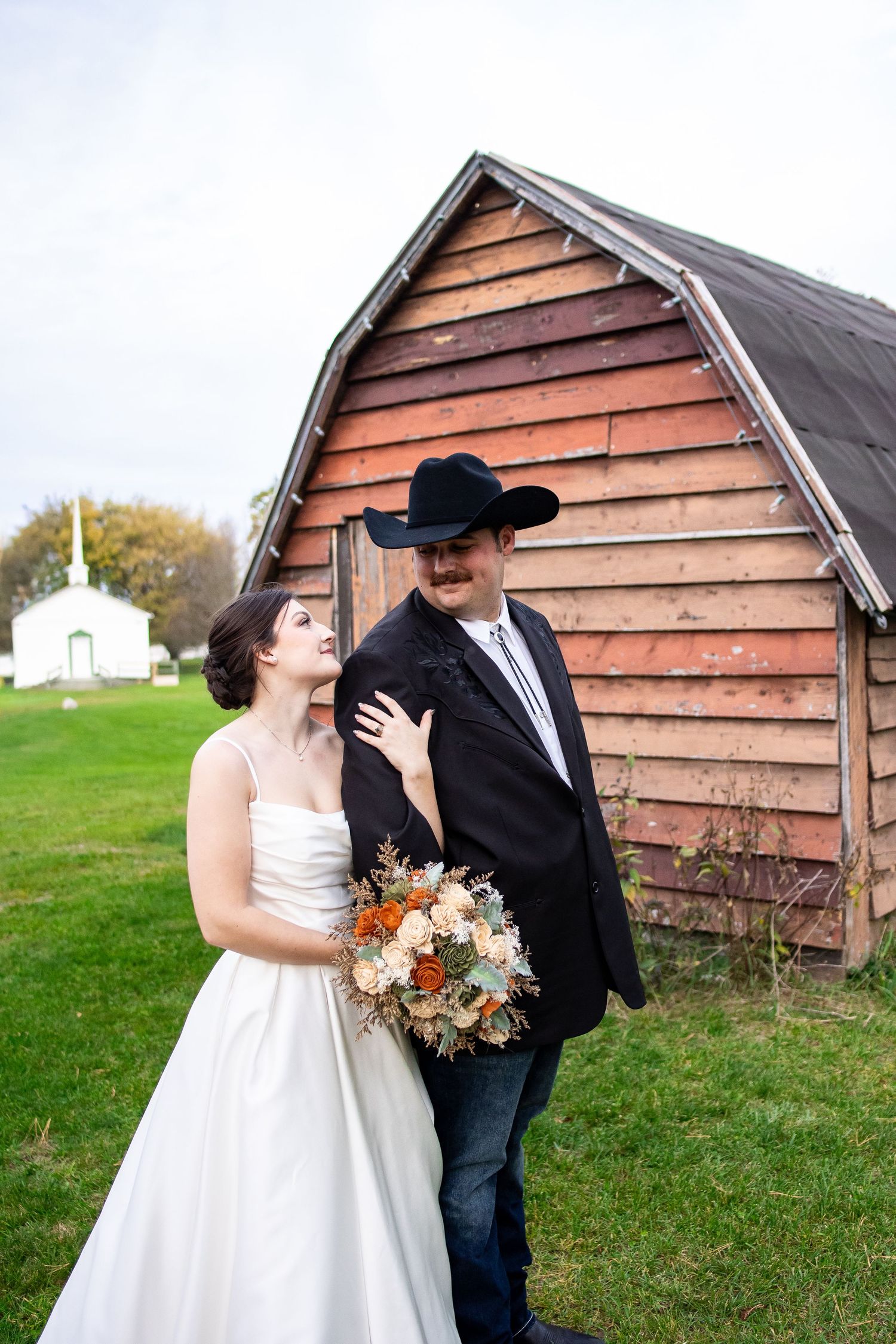 Bride in a white wedding gown holding an autumn bouquet gazes at her groom in a black cowboy hat beside a rustic barn.