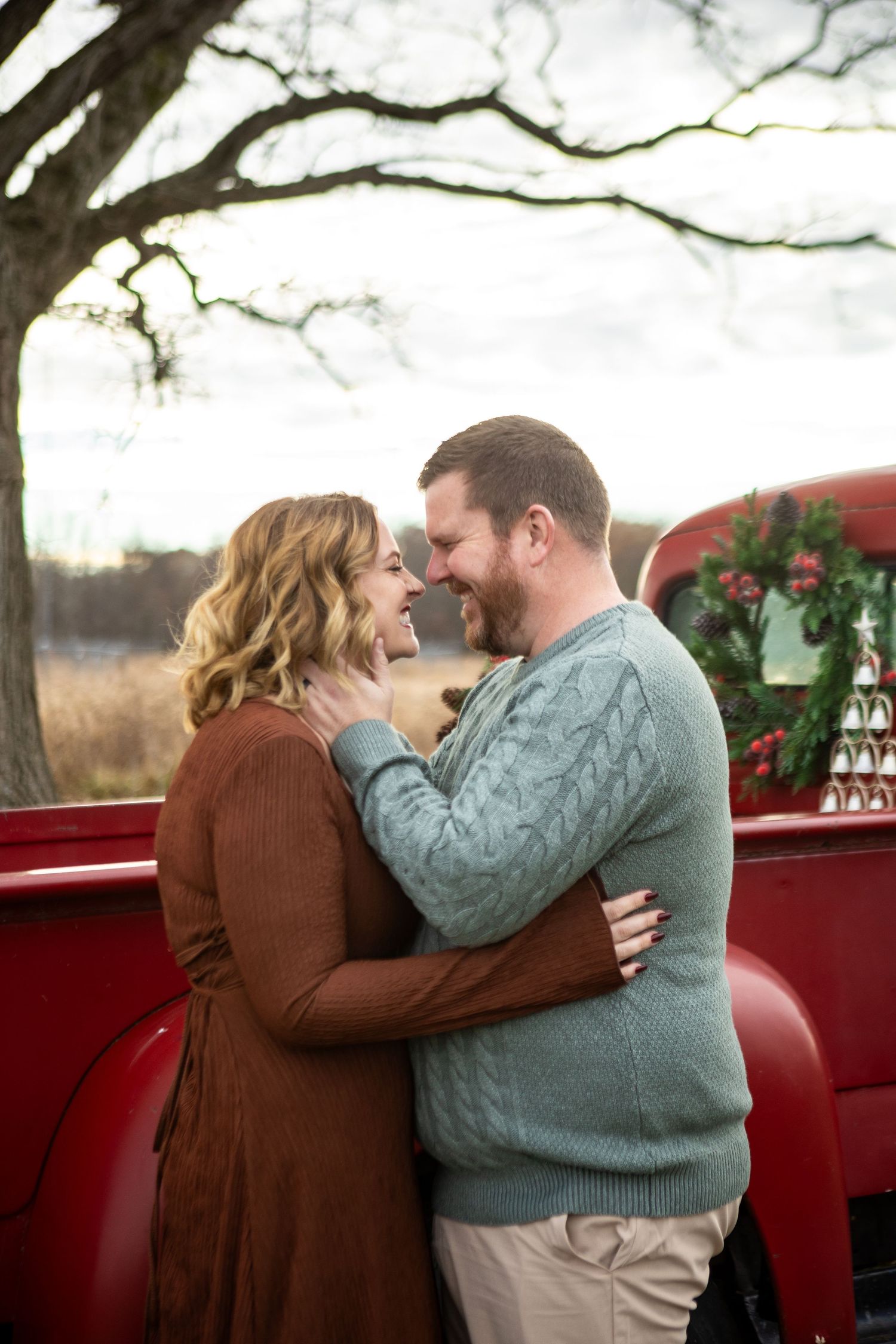A romantic couple embraces in front of a red pickup truck during a winter photoshoot with bare tree branches overhead.