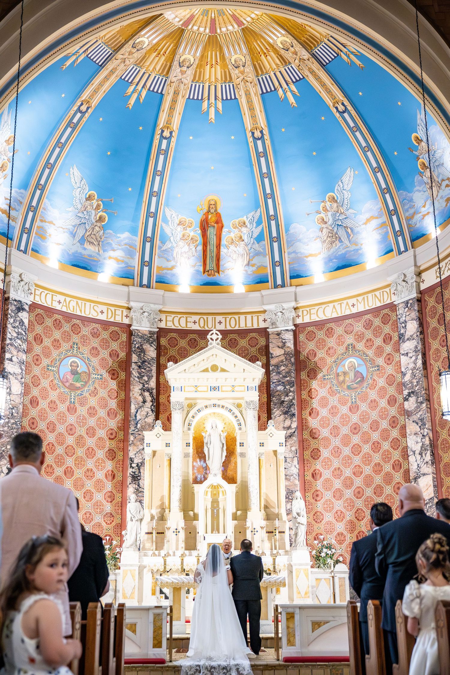 A bride and groom stand at the ornate altar of a stunning Catholic church with a blue dome and golden murals during their wedding ceremony.