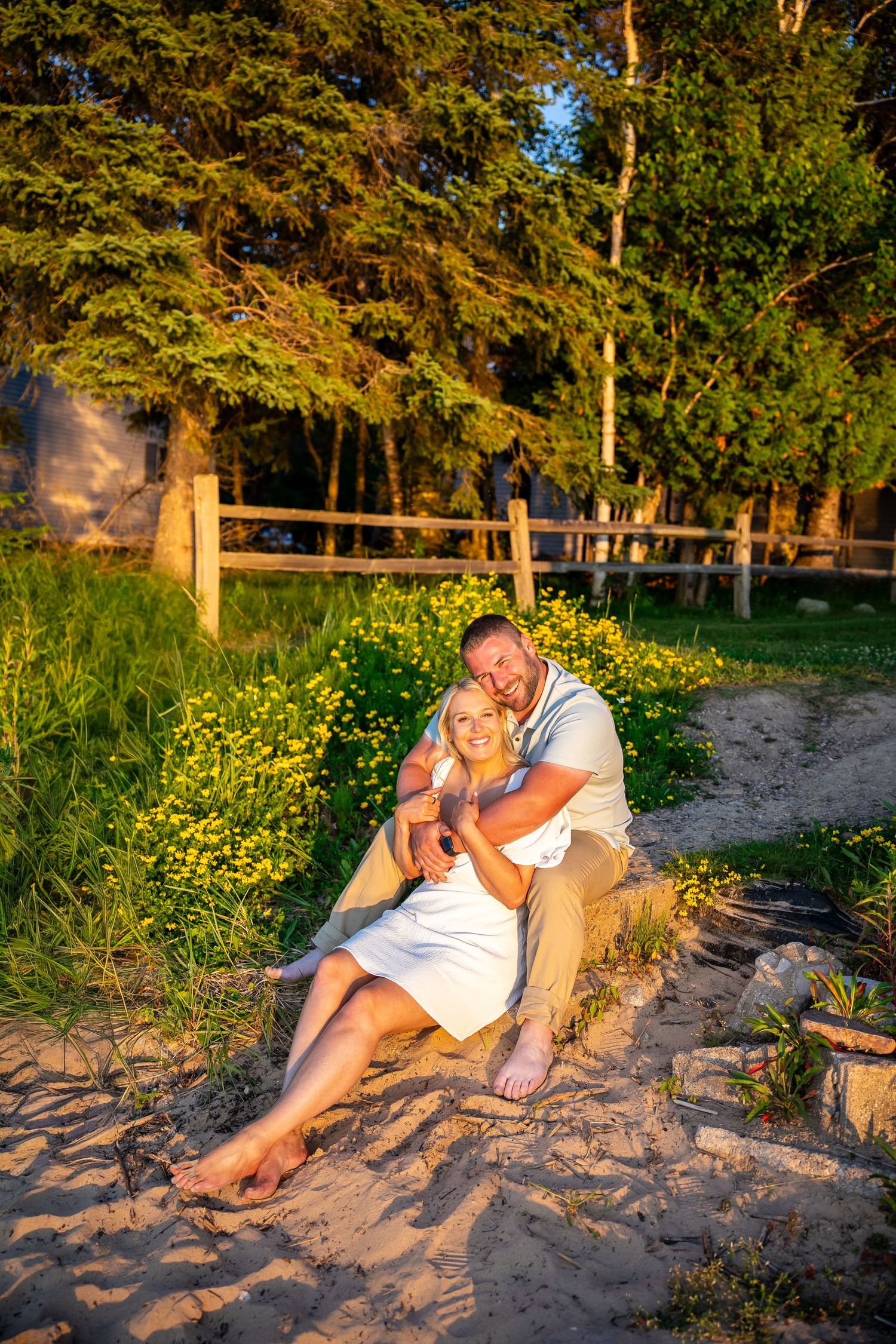 Couple sitting together on sandy beach at golden hour, surrounded by wildflowers and lush green trees.
