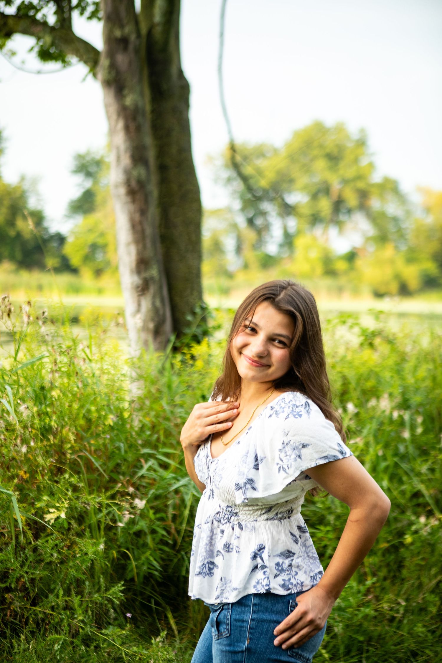 A person in a white floral top and denim shorts poses playfully in a sunny field with green foliage.