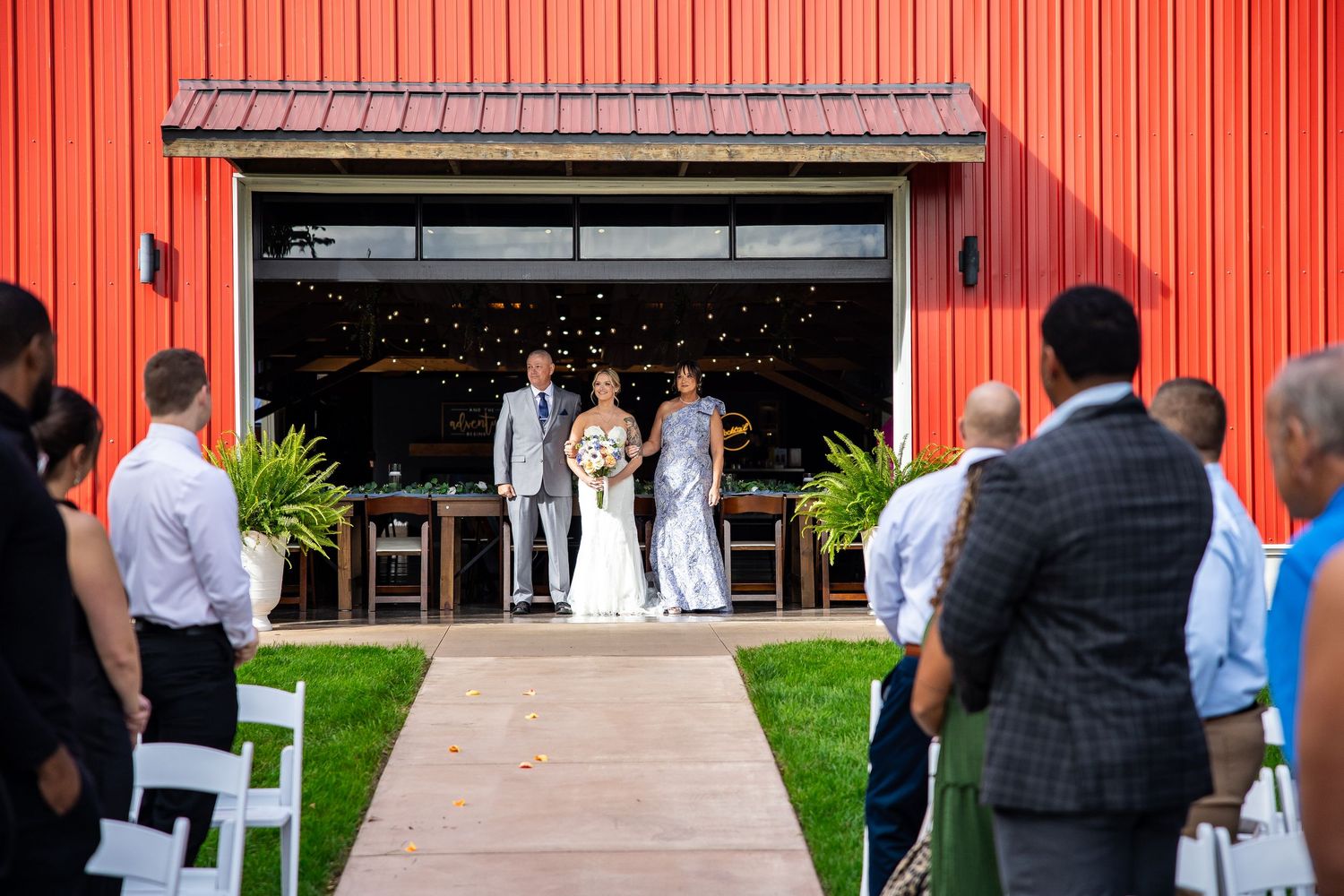 Bride in a white gown walks down the aisle with her father toward the groom as wedding guests watch outside a red barn venue.