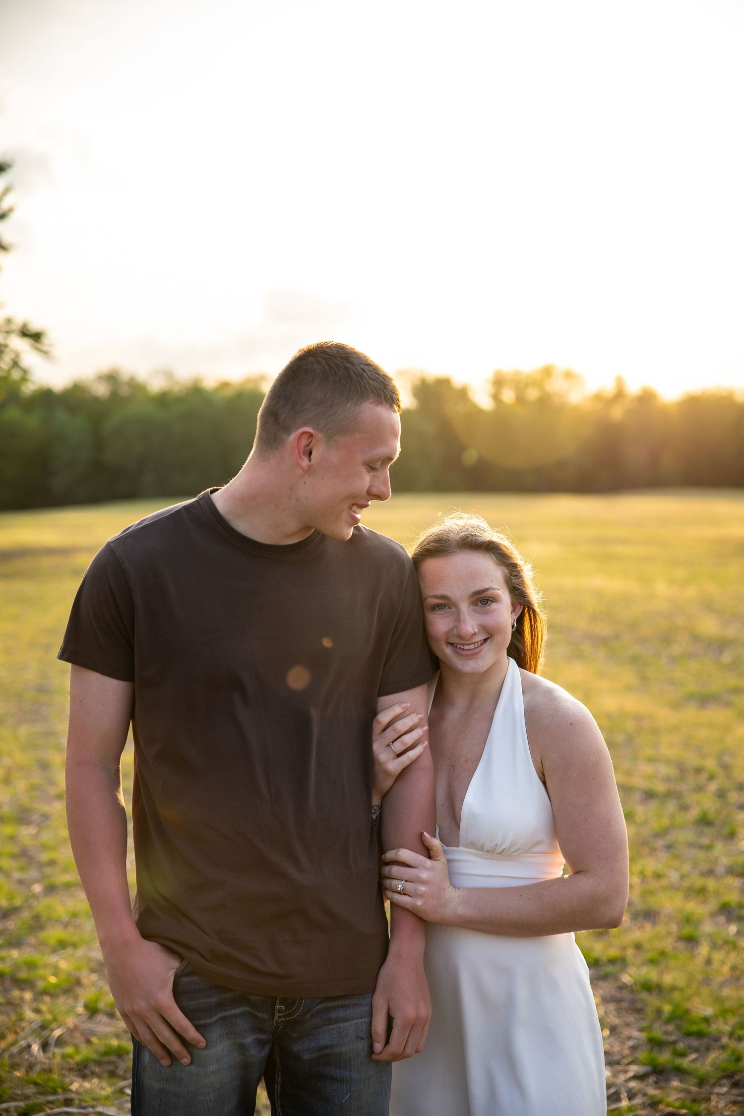 Couple embracing in a sunlit field at golden hour, woman in white dress smiling, man in brown shirt looking at her.