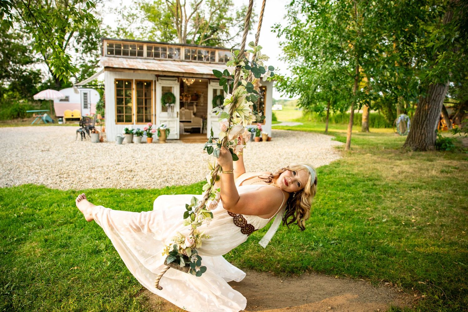 Bride in white gown swinging on floral swing outdoors near rustic cottage surrounded by lush green trees.