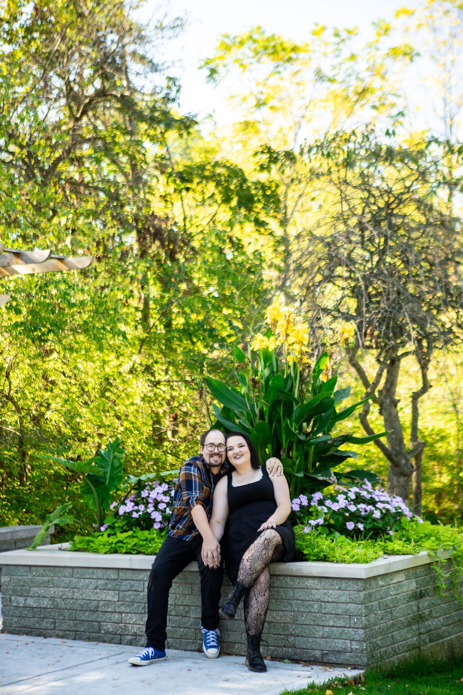 Two people sitting together on a stone wall in a lush garden with green foliage and trees in the background.