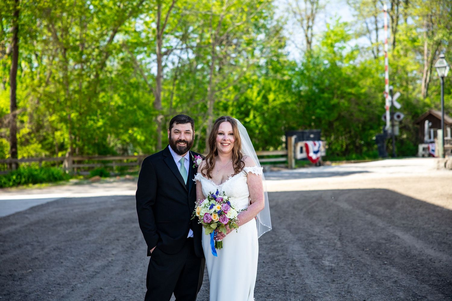 Bride and groom pose outdoors on gravel path surrounded by lush green trees on their wedding day.