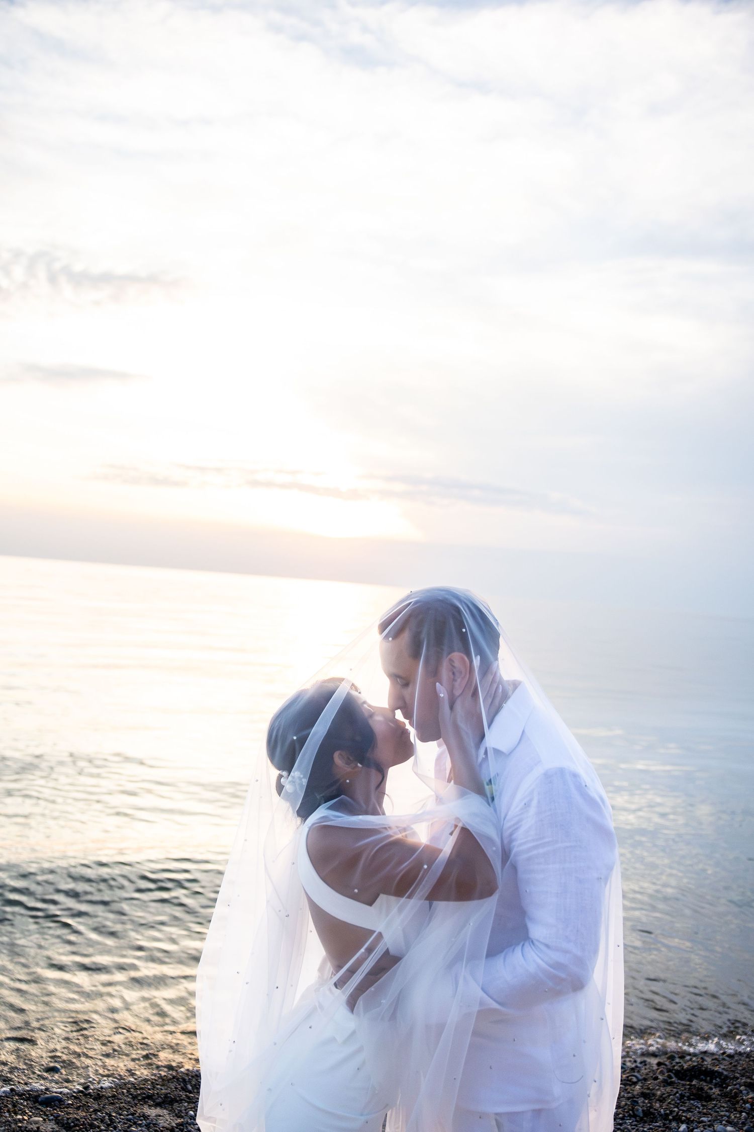 Bride and groom kissing under a veil at sunset by the ocean on their wedding day.