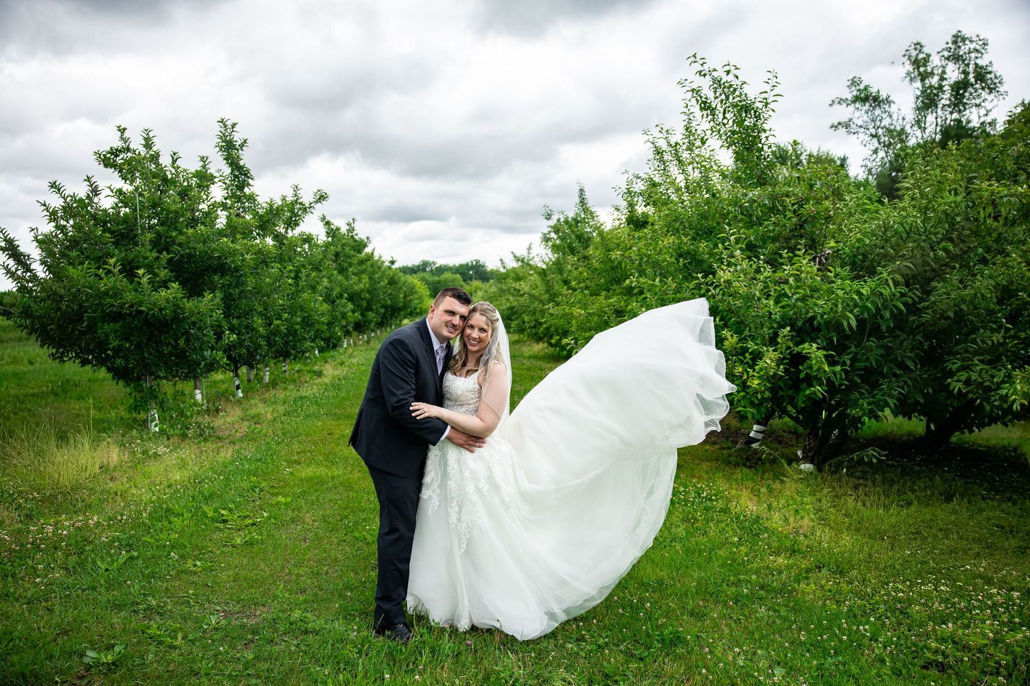 Bride and groom embrace in lush green orchard, wedding veil flowing in wind on cloudy day.
