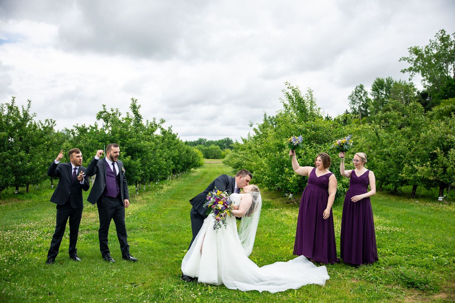 Bride and groom share a kiss surrounded by wedding party in purple attire in a lush green orchard.