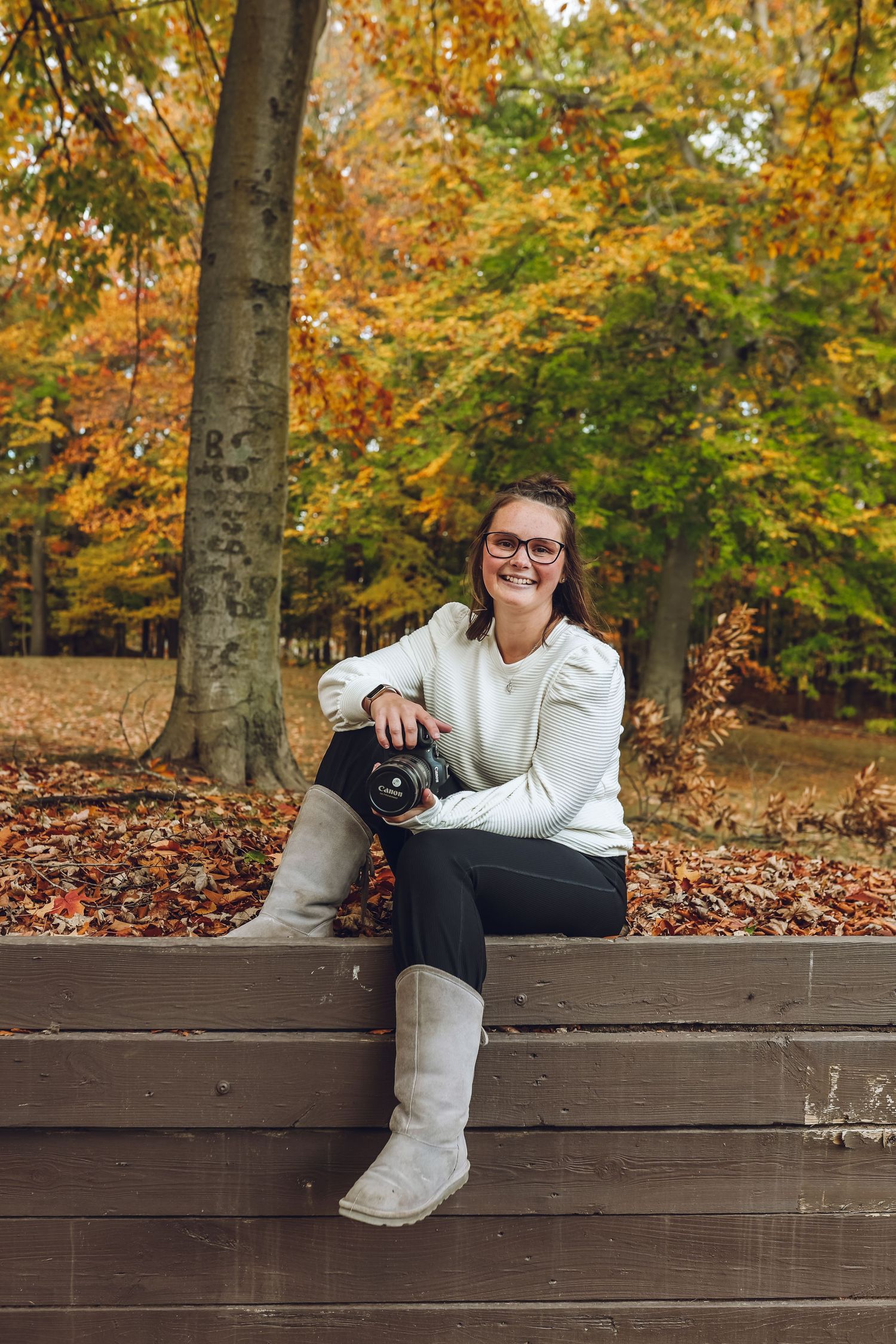 Someone in a white sweater sits with a black dog on a wooden bench surrounded by autumn foliage in a park.