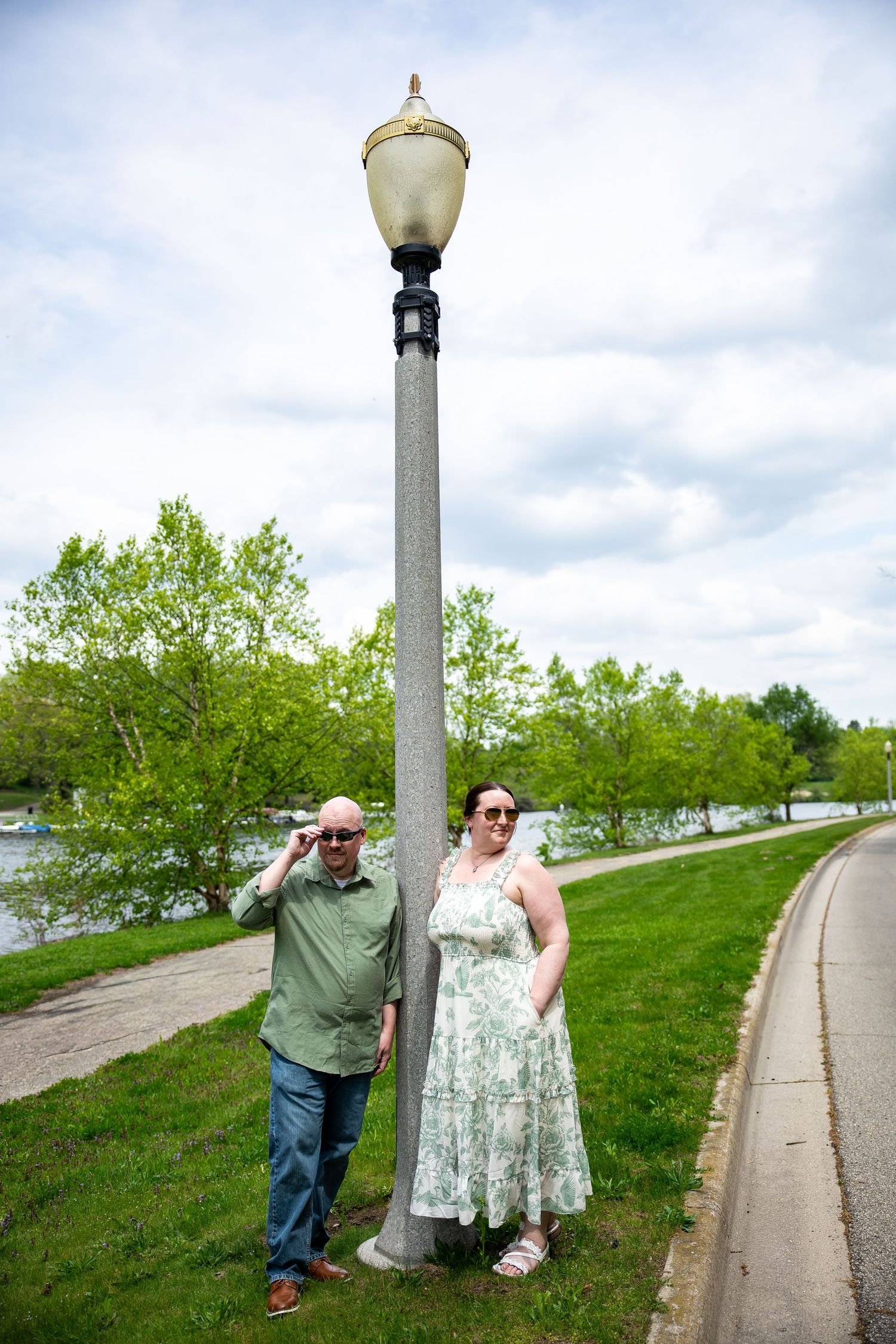 A couple stands by a vintage lamppost in a lush green park, man in green shirt and woman in floral dress.