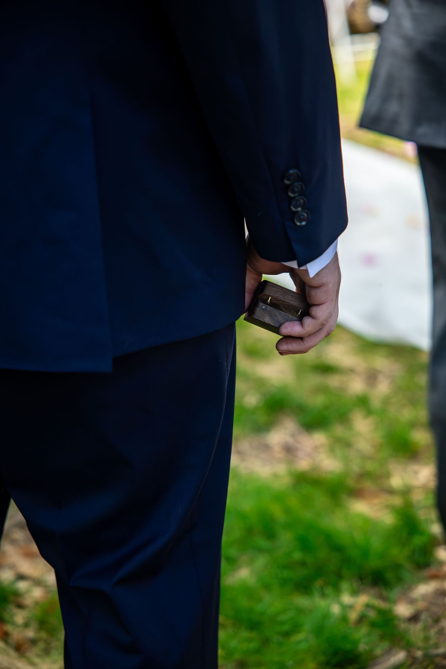 Man in navy suit holding a small ring box behind his back outdoors on green grass at a wedding proposal.